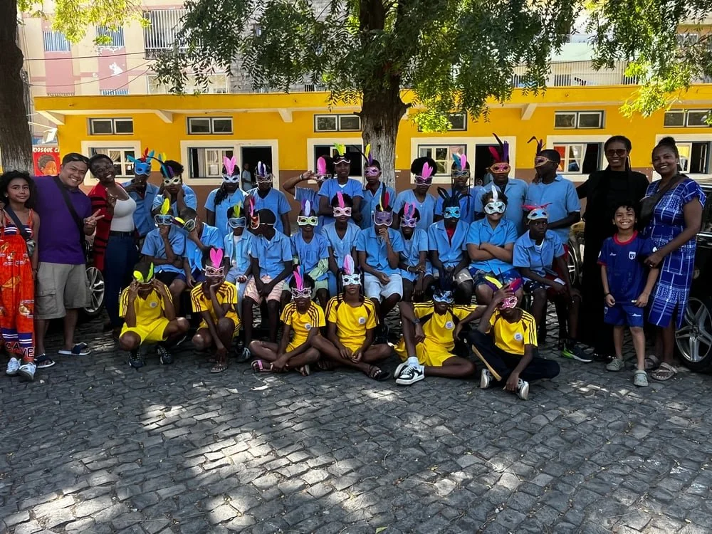 Group of children wearing colorful animal masks, mostly rabbits, posing outdoors in front of a yellow building with adults and a large tree.