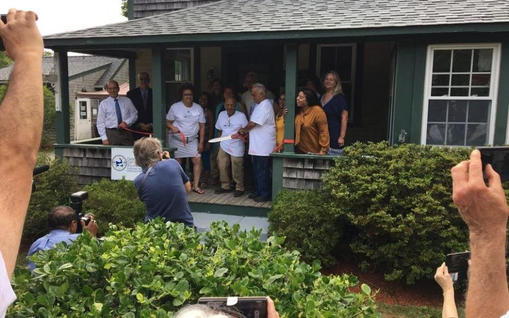 People attending and taking photos of a ribbon-cutting ceremony in front of a house, with some holding cameras and phones, on a porch with a green exterior.