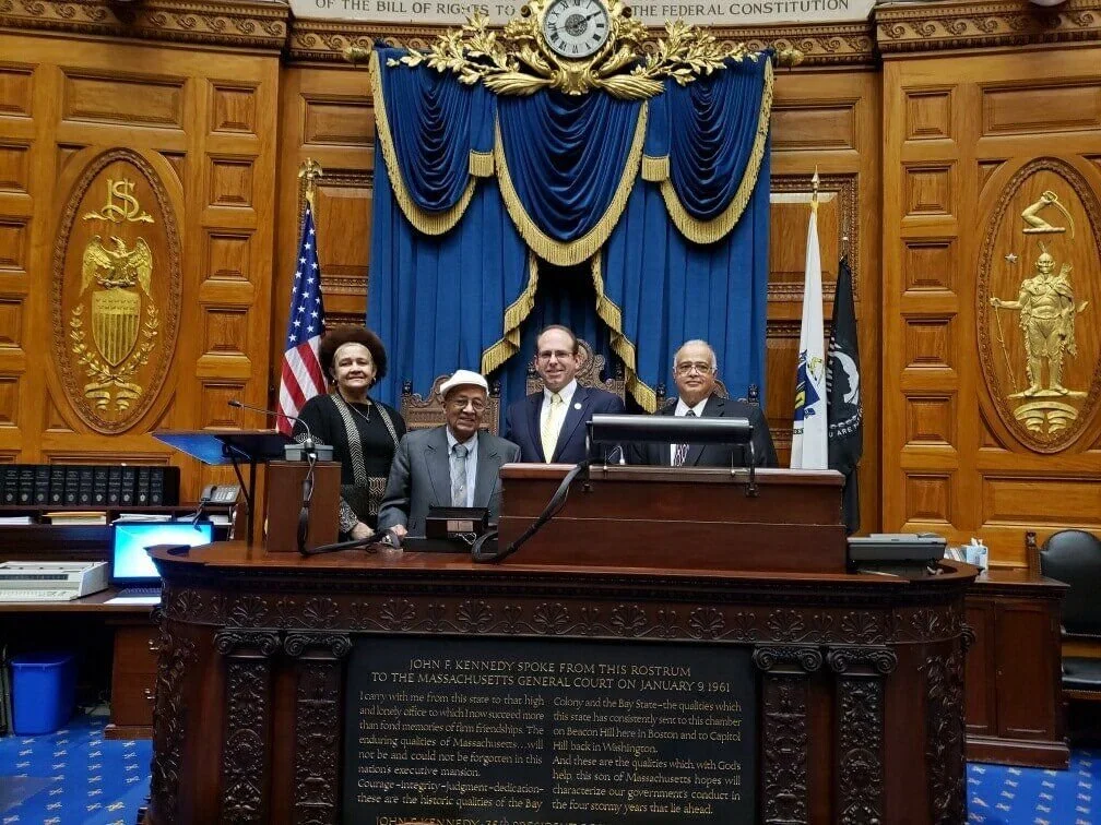 Four people standing in a courtroom with wood-paneled walls, blue curtains, and gold accents. The group includes a woman and three men, one seated and three standing behind. There are American and other state flags. A clock and a bust of John F. Kenn