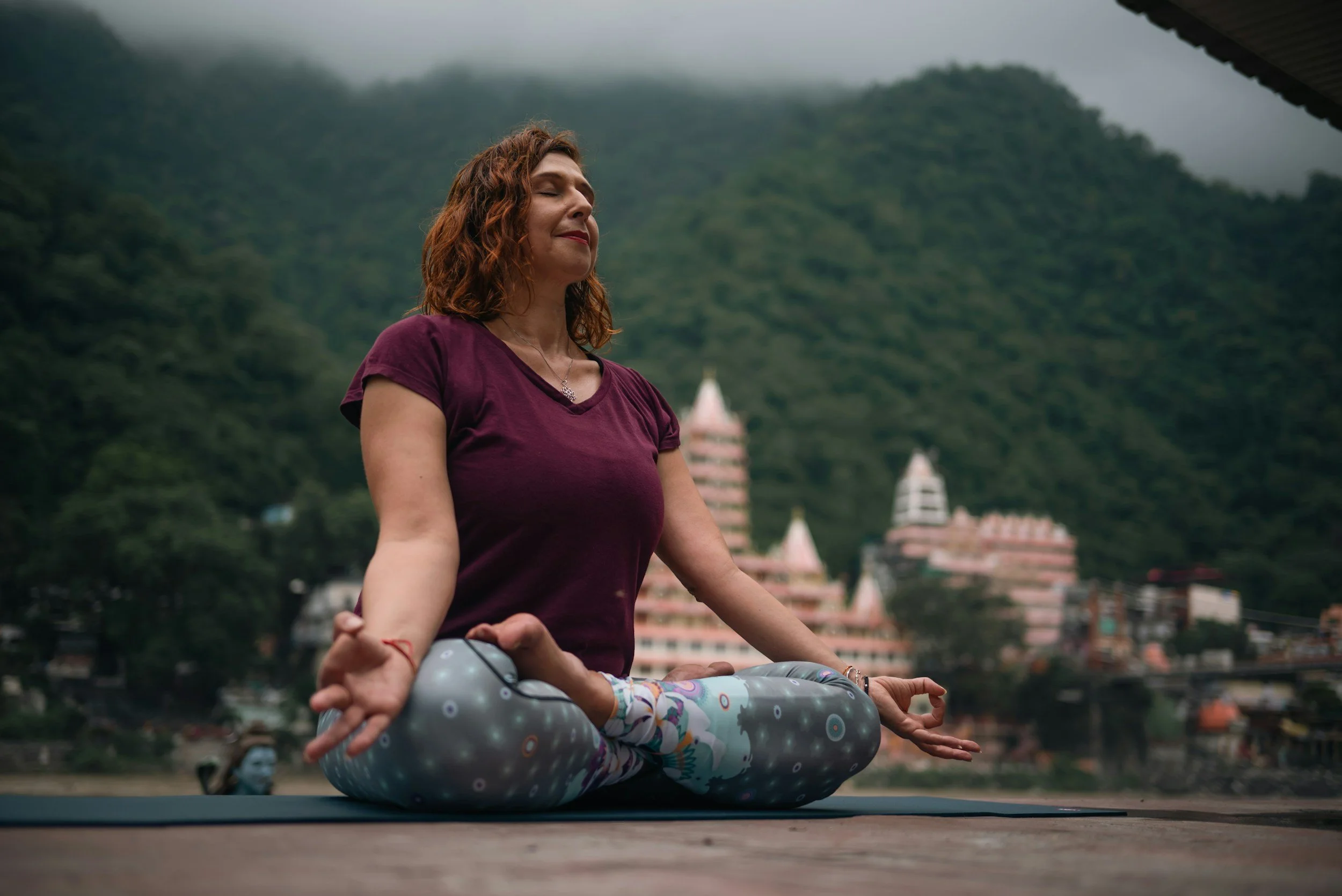 A woman practicing yoga outdoors, seated in a meditative pose with her legs crossed and hands resting on her knees, against a backdrop of a temple and lush green hills.