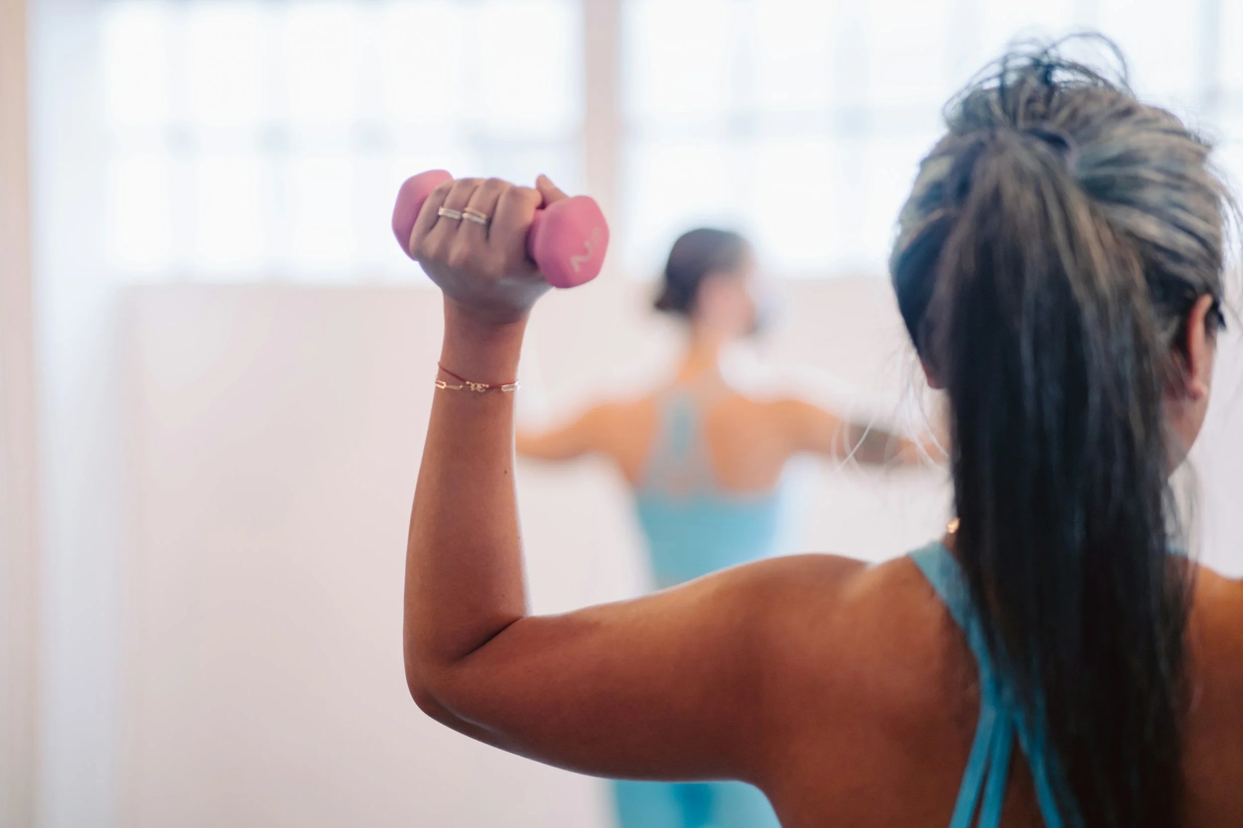 A woman with blonde hair tied back, wearing a blue tank top, holds a small pink dumbbell in her right hand while flexing her arm in a fitness class. In the background, another woman with short dark hair, also in a fitness class, is visible but blurred.