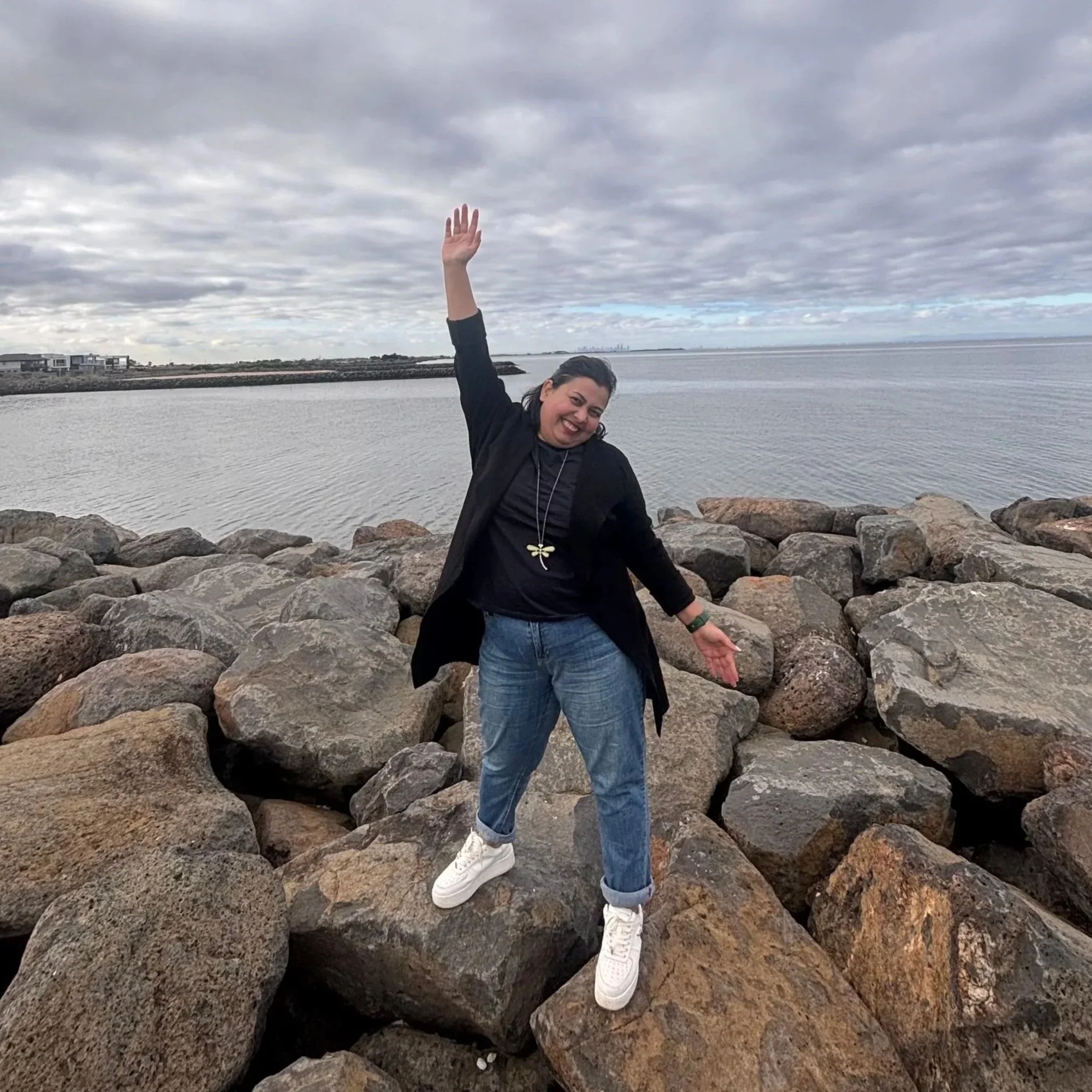 A woman standing on large rocks by the water, smiling and posing with one arm raised, in front of a cloudy sky and calm body of water.