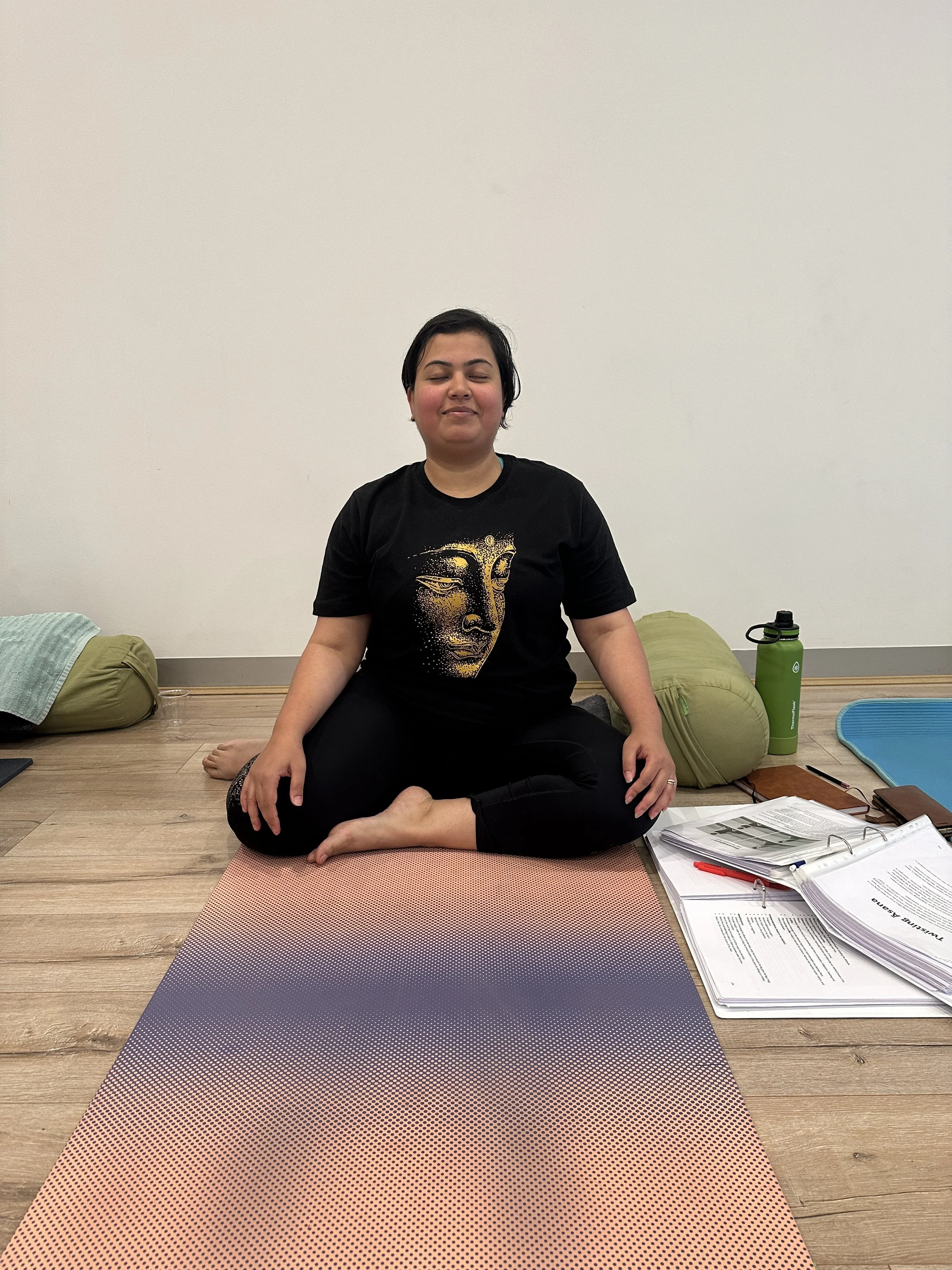 A woman sitting cross-legged on a yoga mat in a room, with her eyes closed and a peaceful expression. She is wearing a black t-shirt with a gold face design, surrounded by yoga props, notebooks, and a water bottle.