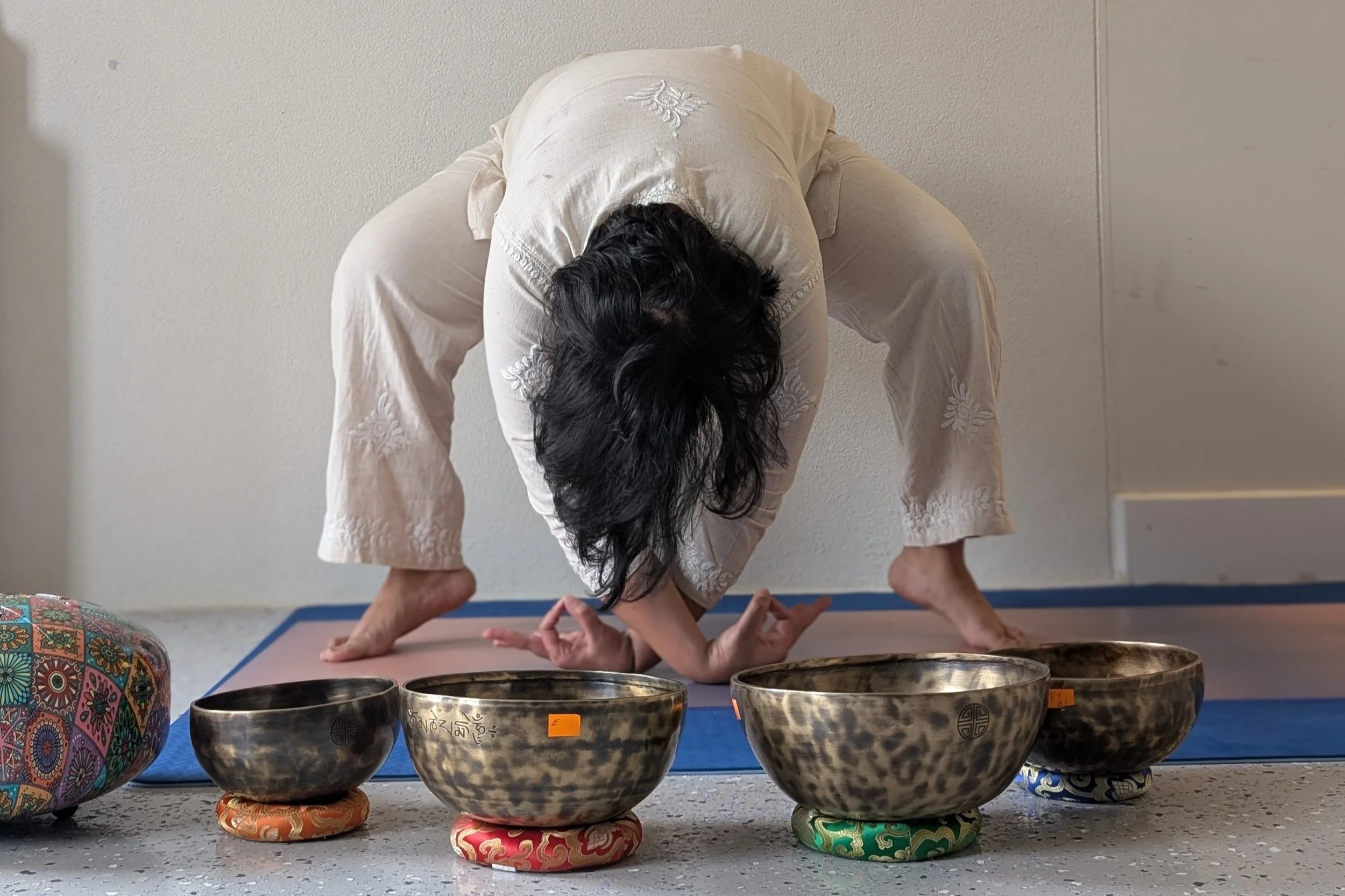 Person with long dark hair doing yoga or meditation pose on a mat, surrounded by Tibetan singing bowls placed on colorful cushions in a bright room.