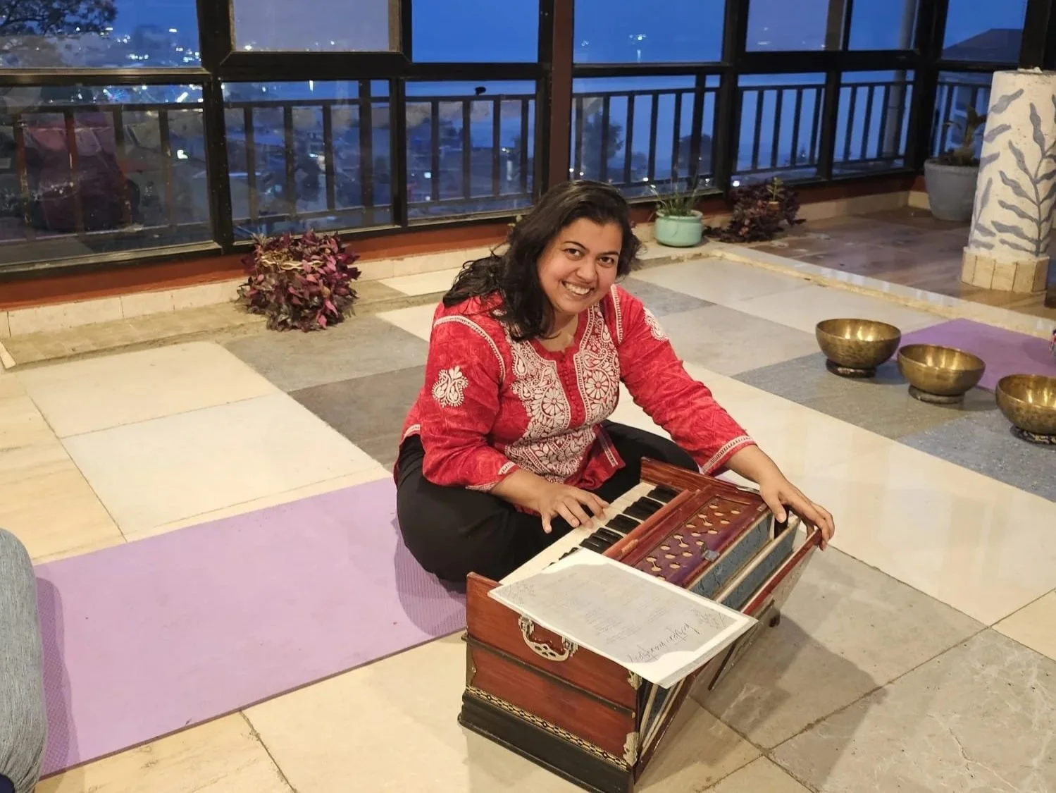 A woman in a red traditional outfit sitting on a purple yoga mat, playing a harmonium, in a room with a tiled floor, potted plants, large windows, and brass singing bowls.