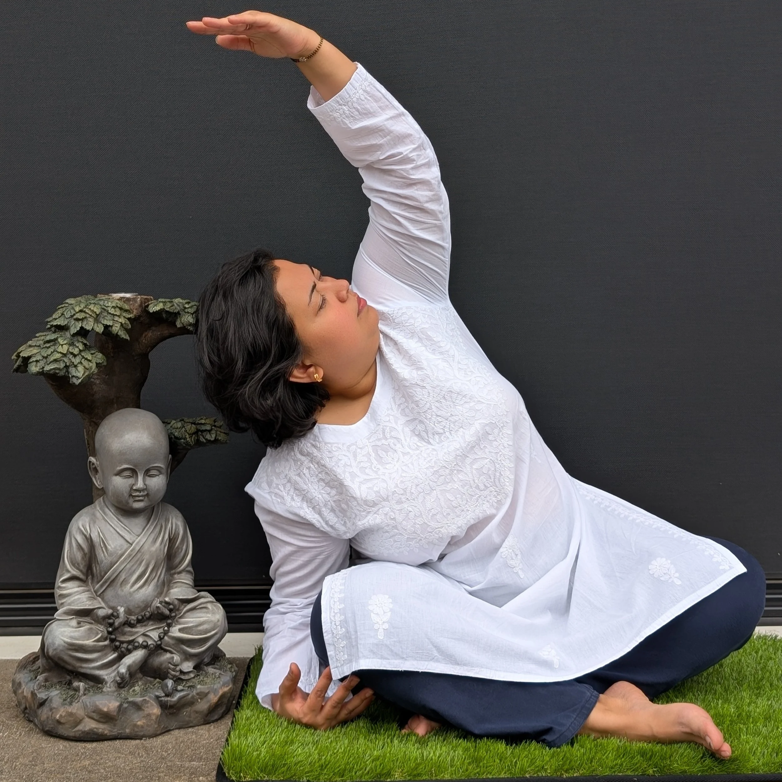 Woman practicing yoga outdoors near a Buddha statue and a decorative tree sculpture, sitting on a grass mat against a black wall.