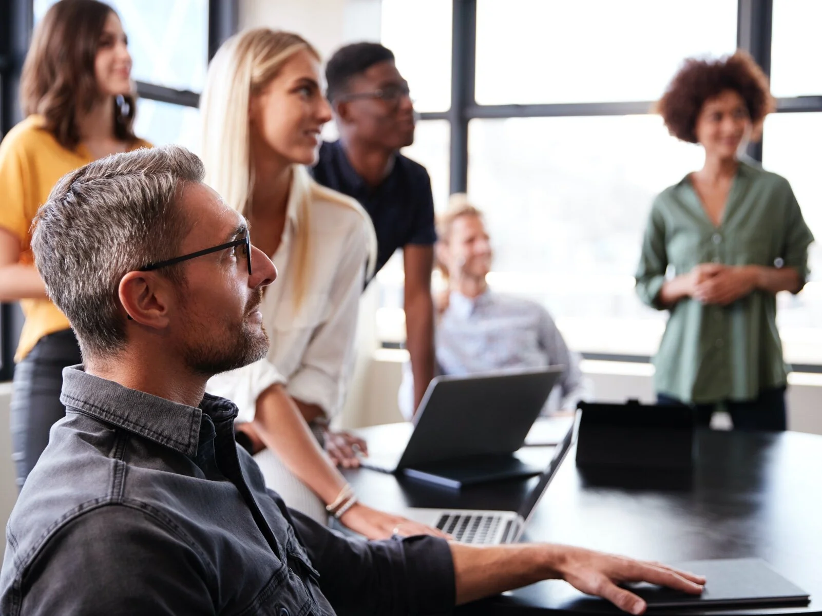 Group of diverse people in a business meeting room, some standing and some seated, with large windows in the background.