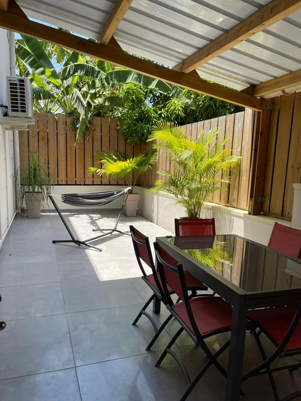 Patio extérieur couvert à Mayotte avec table et chaise, plantes vertes en pot, clôture en bois, et un sèche-linge suspendu, avec des plantes tropicales en arrière-plan. Services de conciergerie, location courte durée. Les concierges Mayotte.