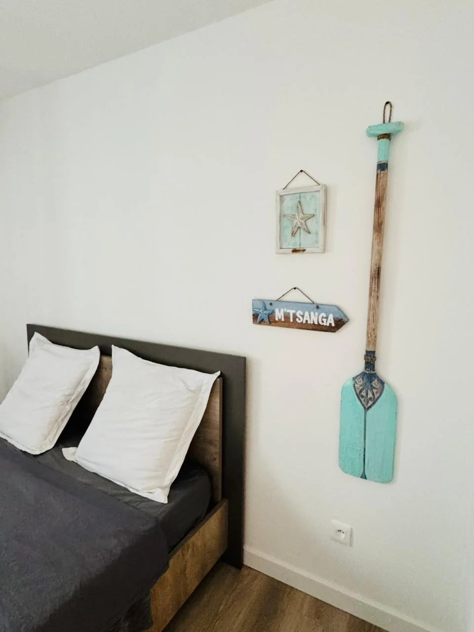 Location de style côtier avec une peinture en forme de paddle, une pancarte avec le nom M'Tsanga, et un tableau avec une étoile de mer, au-dessus d'un lit avec deux oreillers blancs. Les concierges Mayotte. Cosy été vacances île tropicale Mayotte.