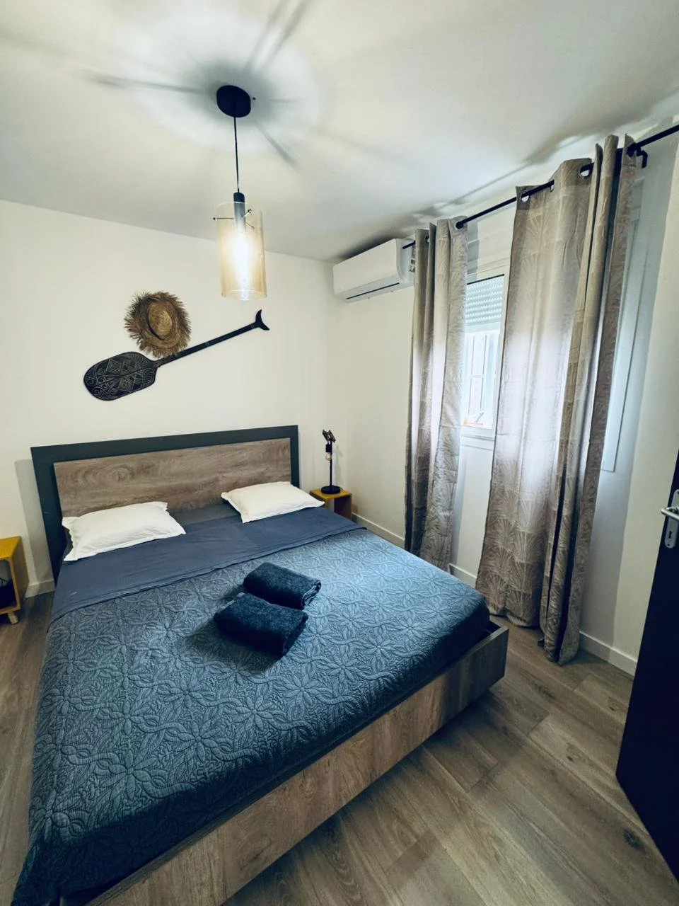Chambre avec un lit double, rideaux, une unité de climatisation, décoration murale de paddle, chapeaux, et une lampe suspendue. Services de conciergerie, location courte durée. Les concierges Mayotte. Cosy été vacances île tropicale Mayotte.