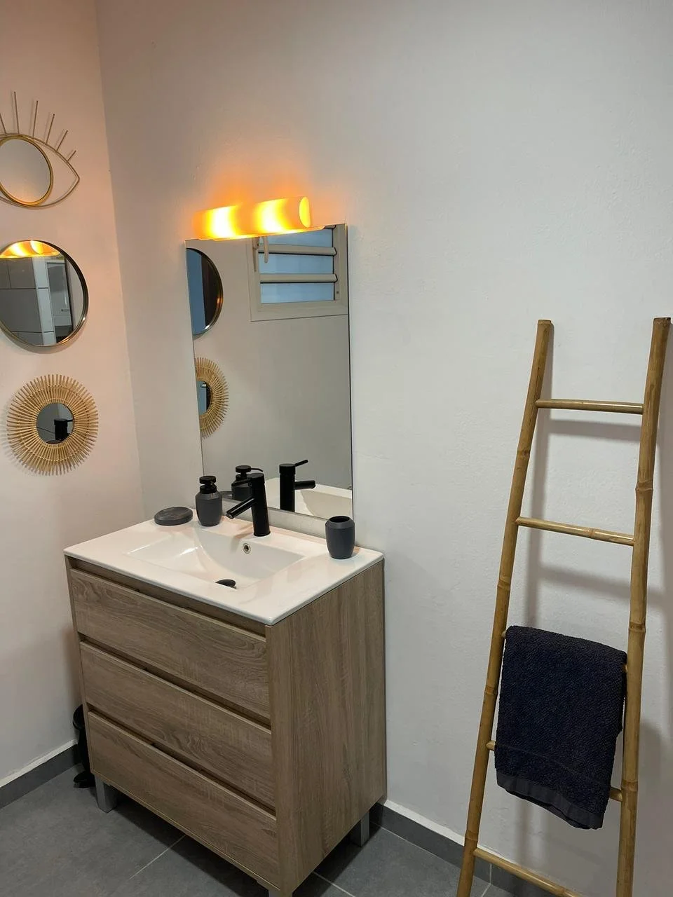 Un lavabo avec un miroir au-dessus, un meuble en bois avec trois tiroirs, des accessoires noirs, un porte-serviette noir, une échelle en bambou, quelques miroirs décoratifs sur le mur, et un éclairage warm. Les concierges Mayotte. Cosy été vacances.