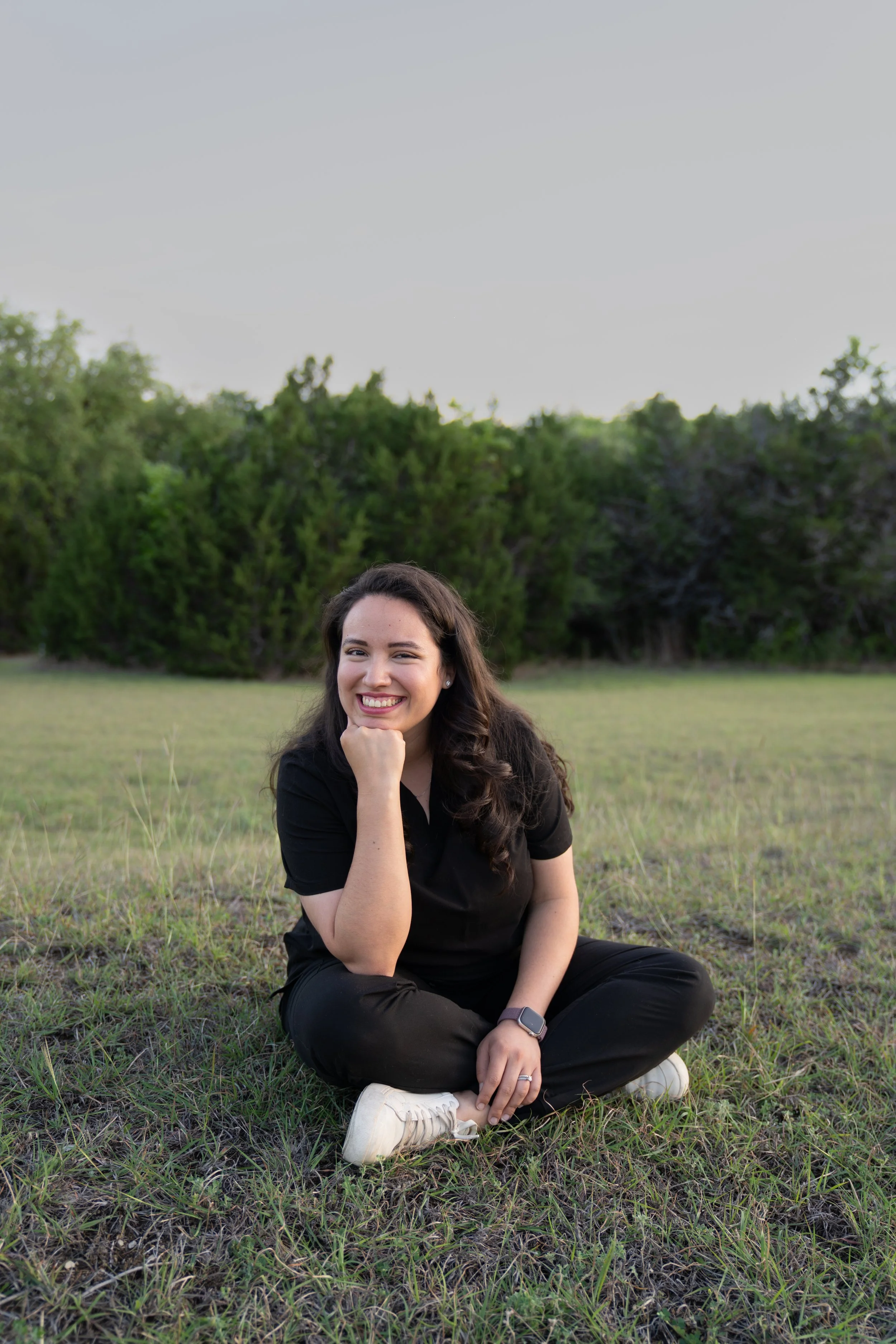 A young woman with long dark hair, smiling, sitting cross-legged on the grass in a field with green trees in the background.