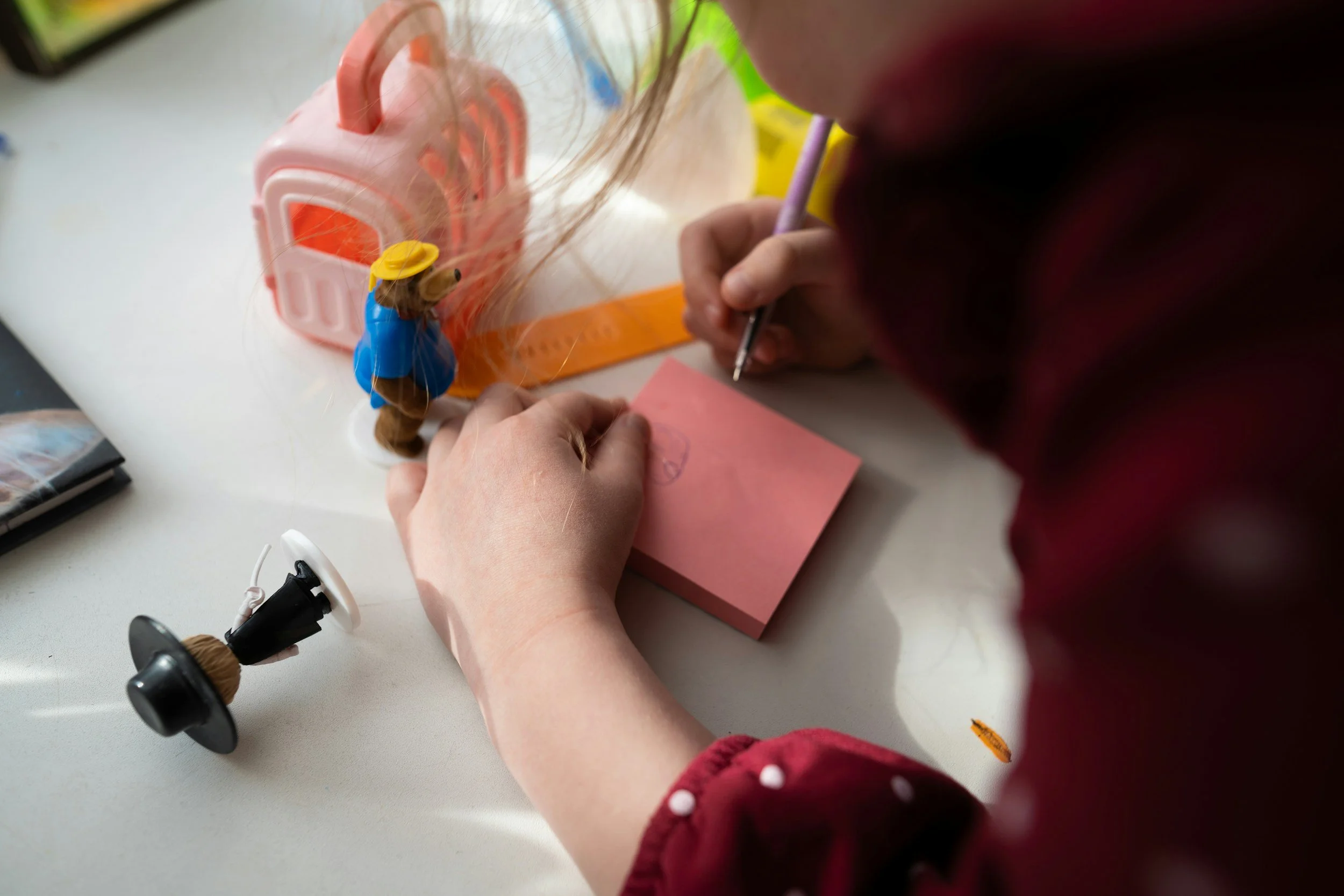 A person is drawing on a pink sticky note, with various objects on a white desk, including a small doll figurine, a pink toy house, and a miniature spool of thread.