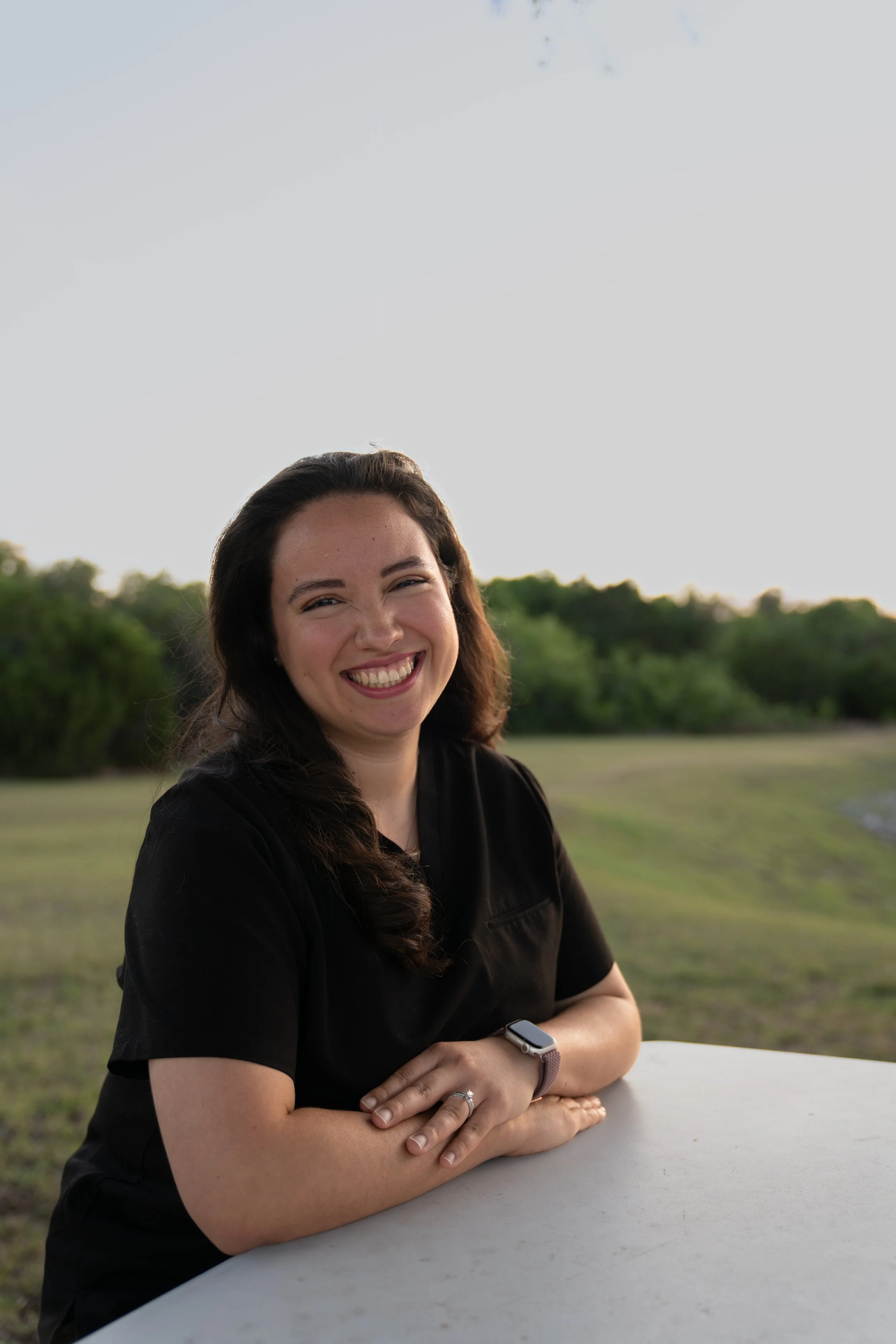 A woman with long dark hair smiling outdoors at sunset, wearing a black shirt and a smartwatch, sitting at a white table with green trees in the background.