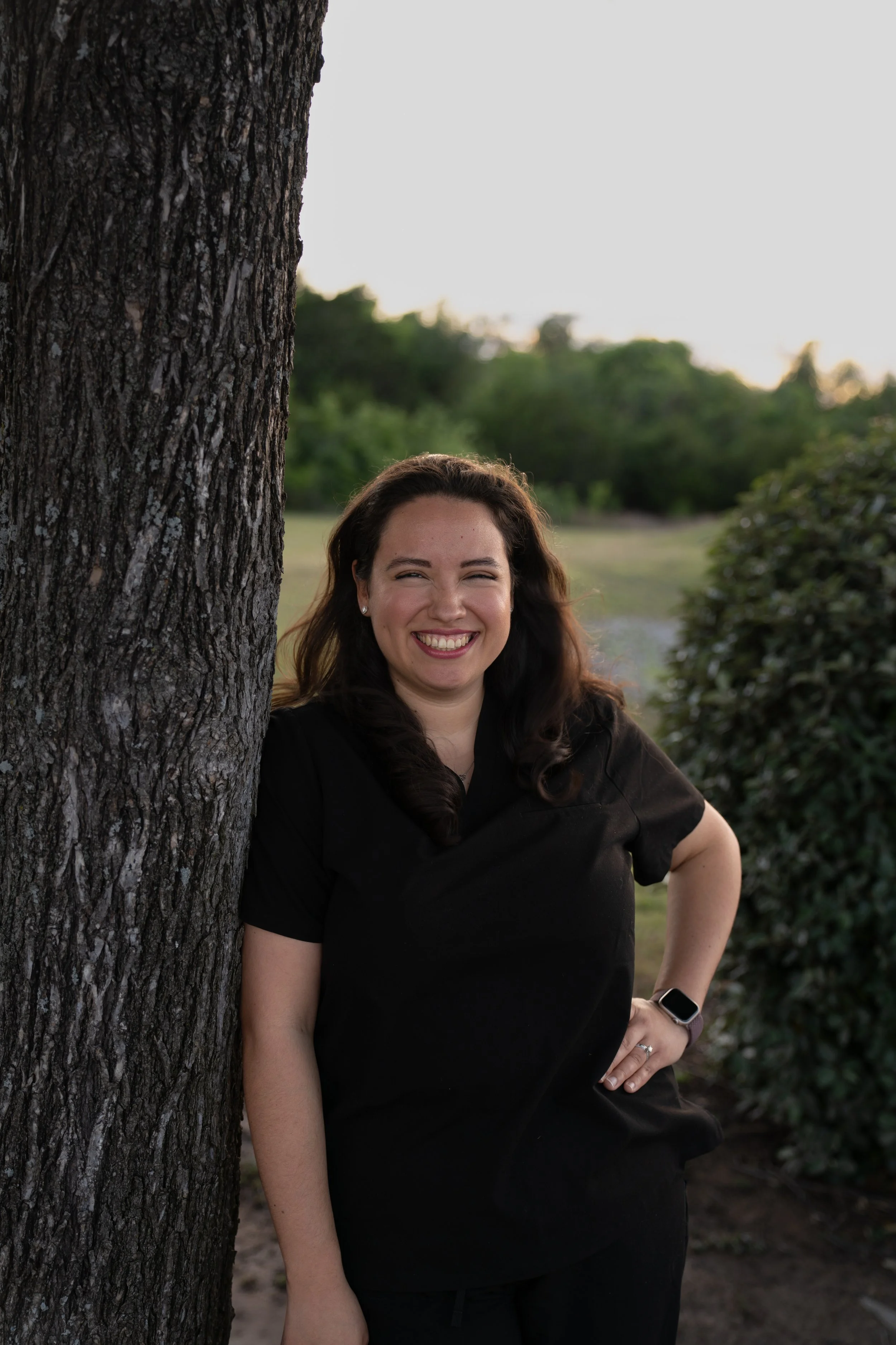 A woman with long brown hair smiling and posing outdoors near a tree during sunset, wearing a black shirt and smartwatch.