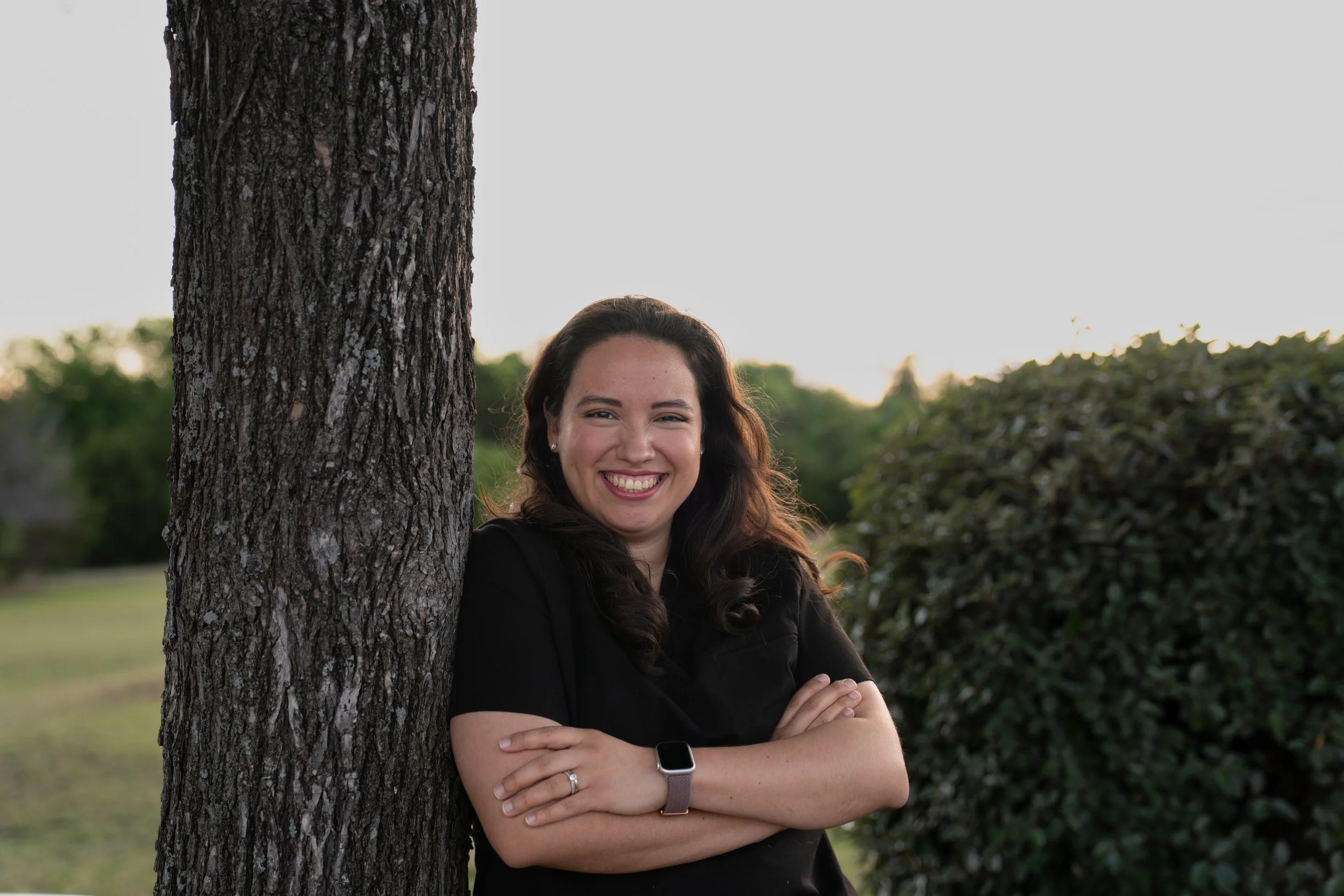 A woman with long dark hair smiling, standing outdoors beside a tree and a bush, wearing a black shirt and a smartwatch, with her arms crossed.
