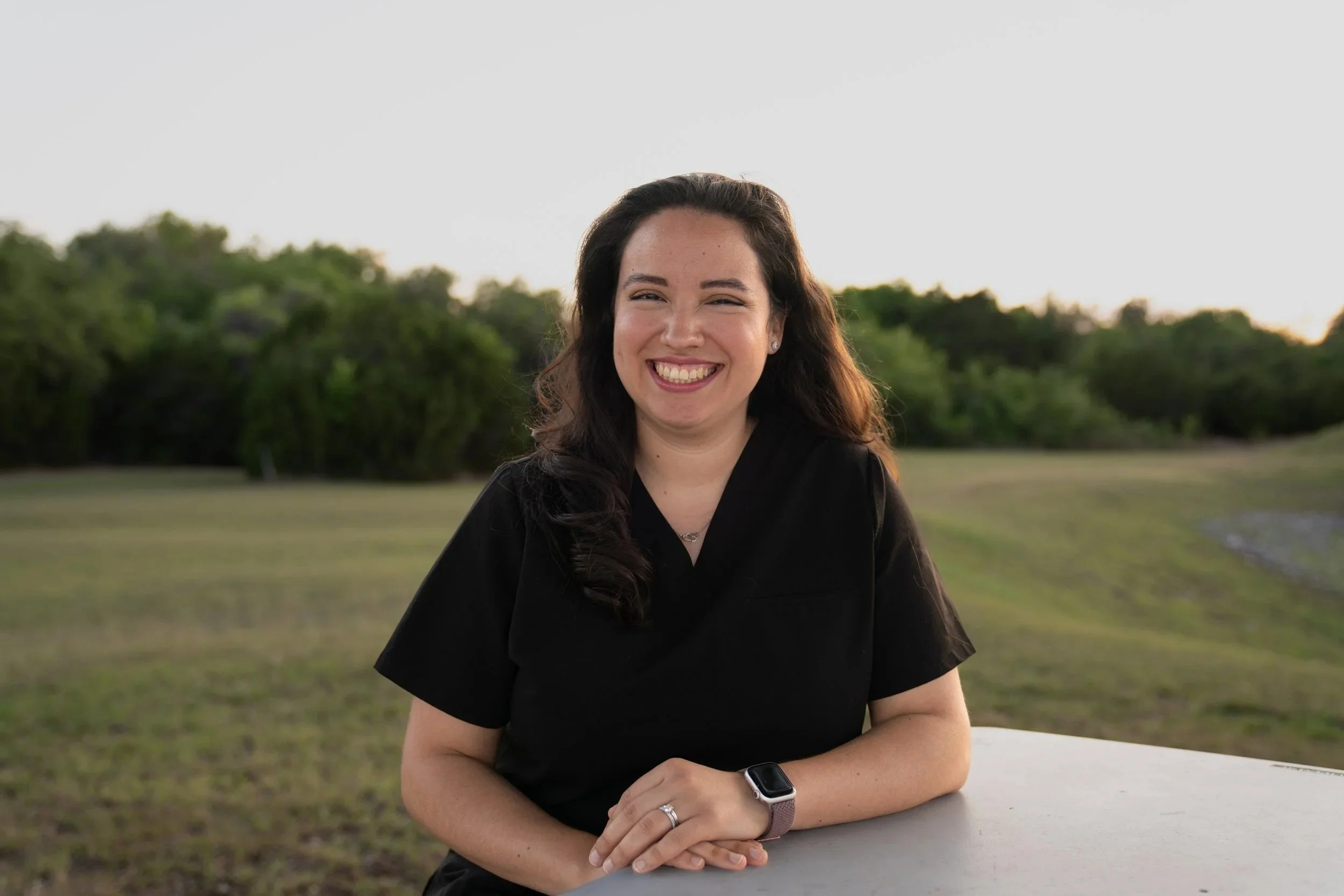 A woman wearing a black shirt smiling outdoors at sunset, leaning on a white surface with a scenic background of grass and trees.