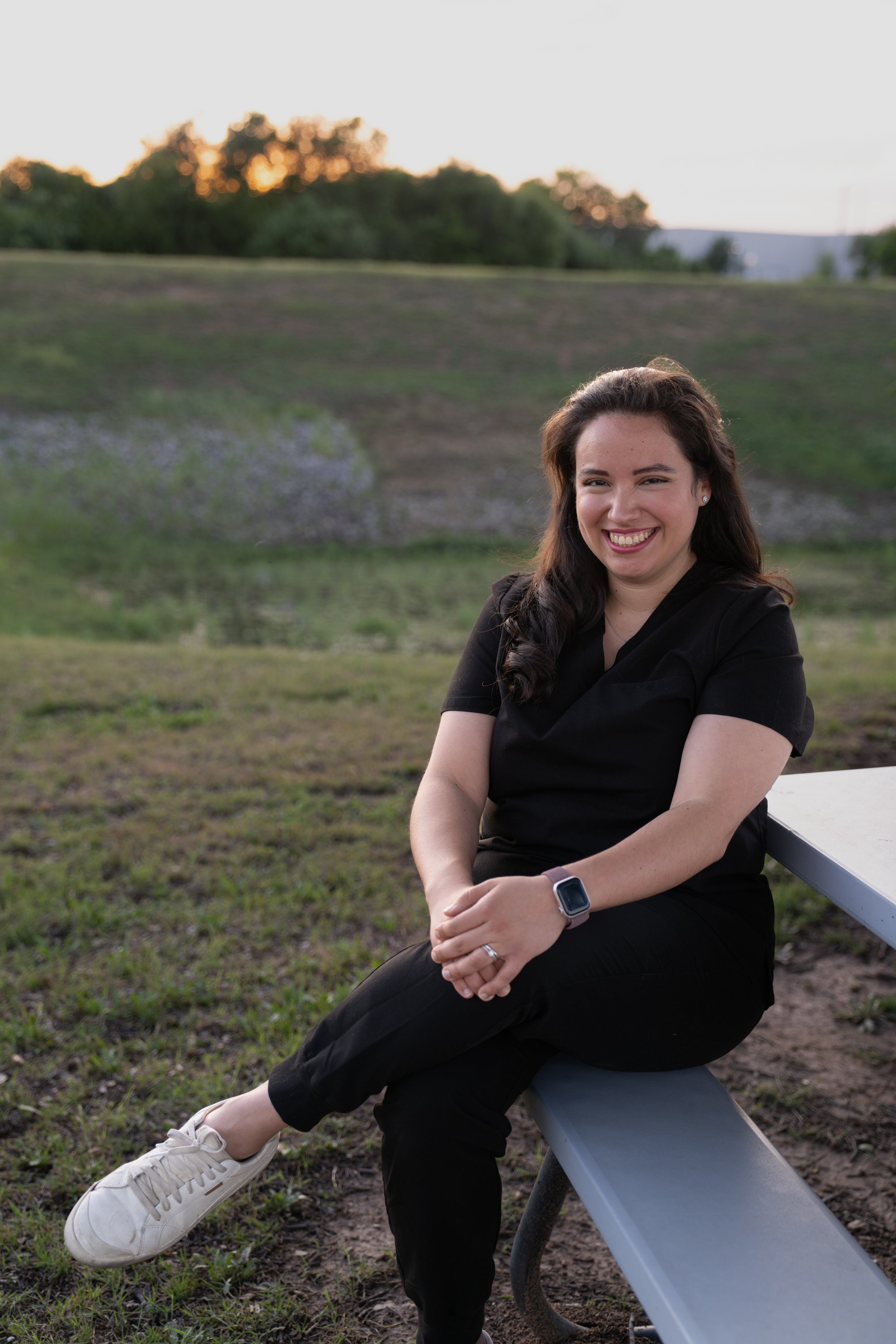 Woman sitting on a bench outdoors, smiling, wearing black scrubs, a smartwatch, and white sneakers, with a grassy field and trees in the background at sunset.