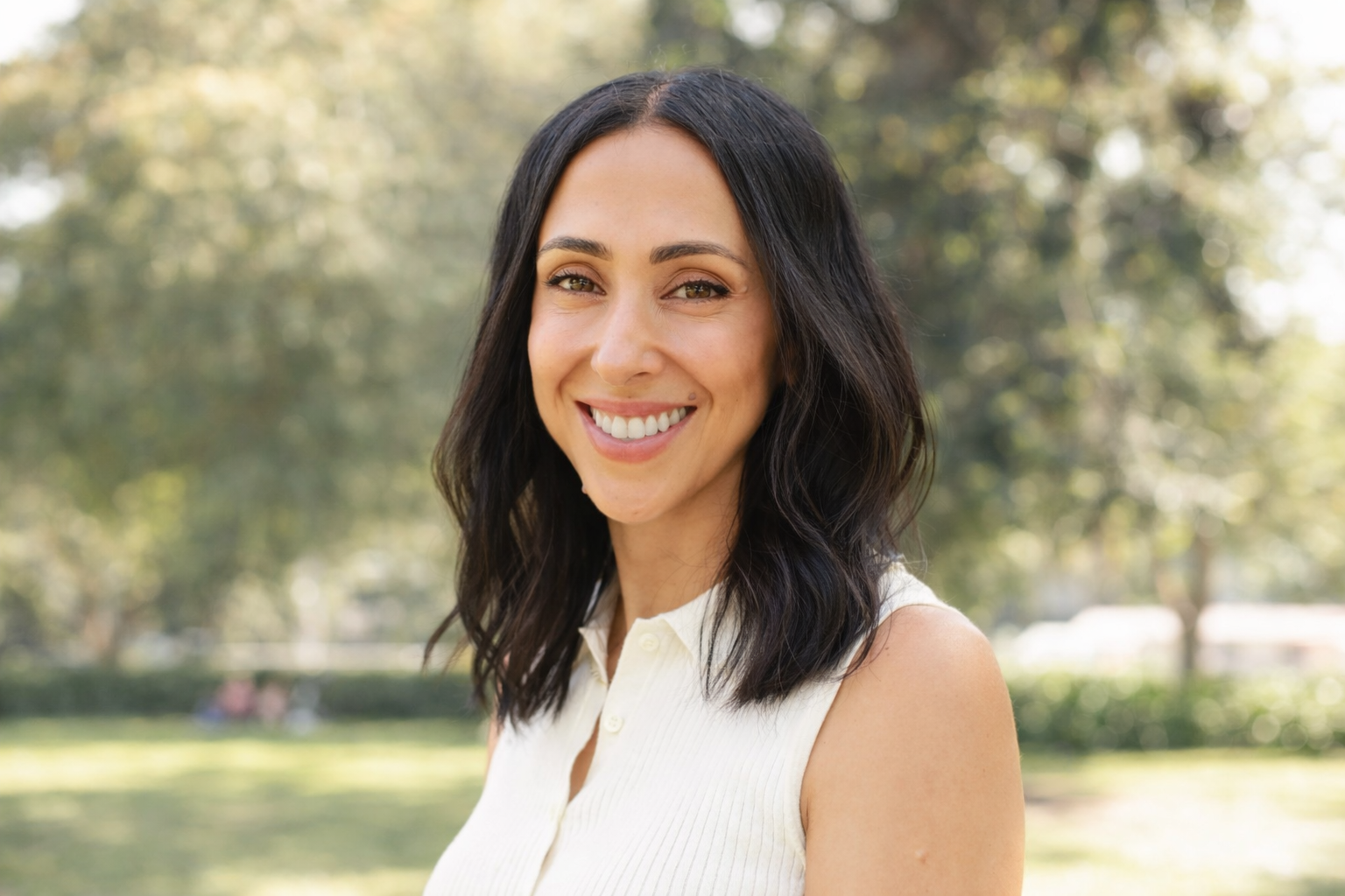 A woman with black, wavy hair and a white sleeveless top smiling outdoors in a park with trees and sunlight in the background.