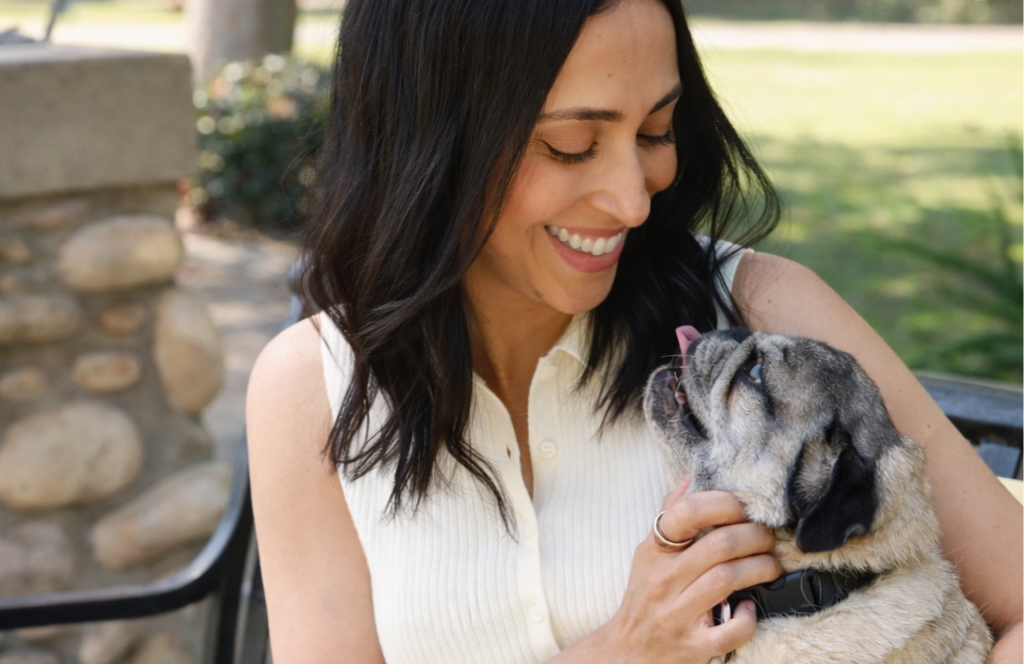 A woman smiling and holding a small pug dog outdoors in a park-like setting.