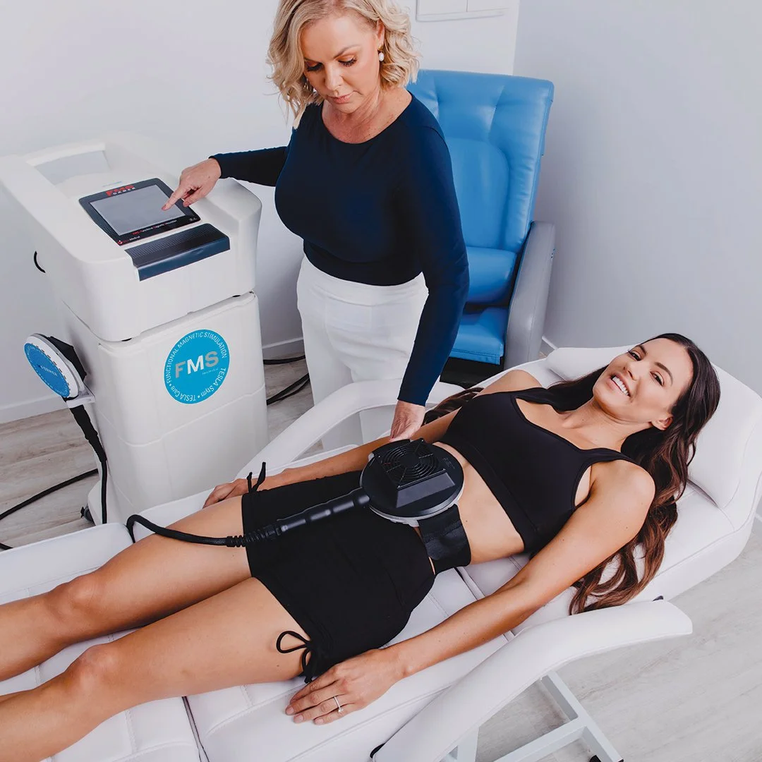 Woman lying on a treatment bed receiving a body contouring procedure with a handheld device, while a technician operates a machine nearby in a clinical setting.