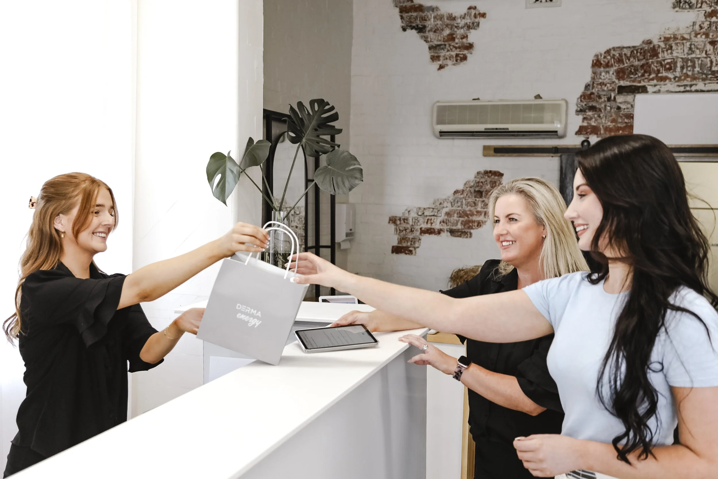 A young woman at a reception desk is handing a shopping bag to a smiling customer, with two other smiling women standing nearby, in a modern interior with brick walls and a white desk.