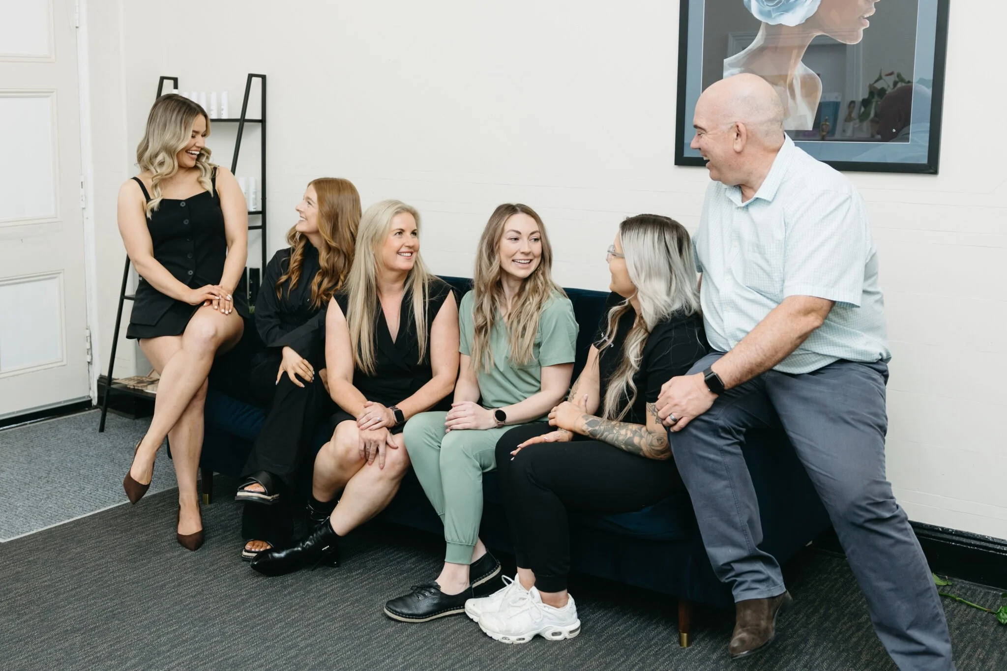 Group of six people sitting and standing in a waiting room, smiling and engaging with each other.