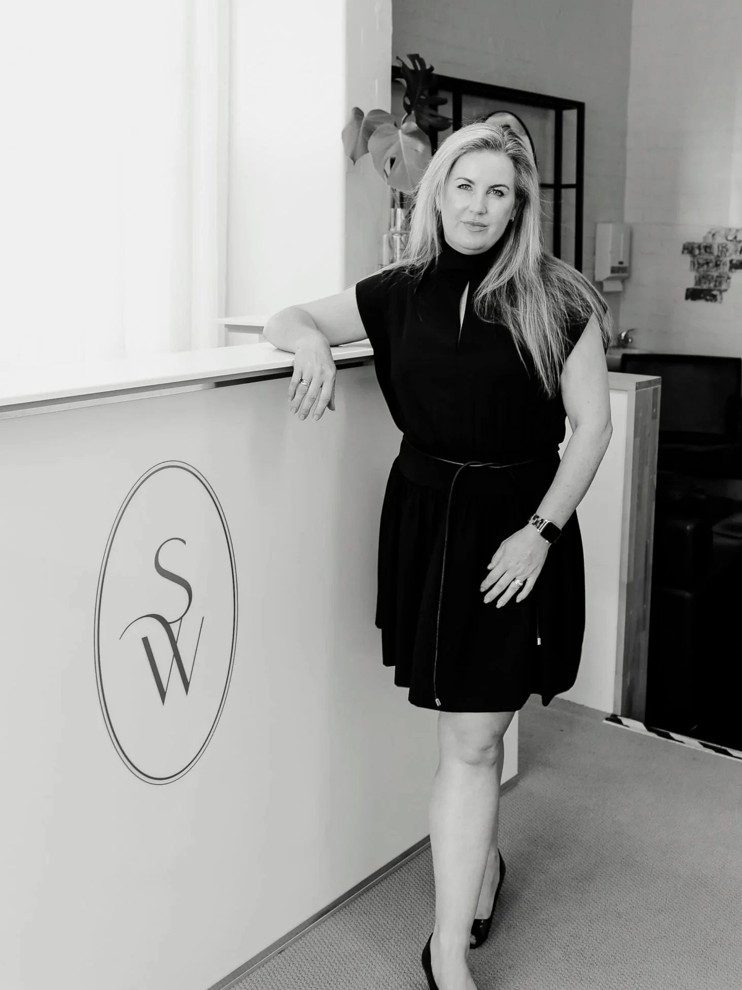 Black and white photo of a woman with long blonde hair wearing a black dress, standing next to a reception desk with a logo that has the letters S and W. She is leaning on the desk with her right arm and smiling at the camera.