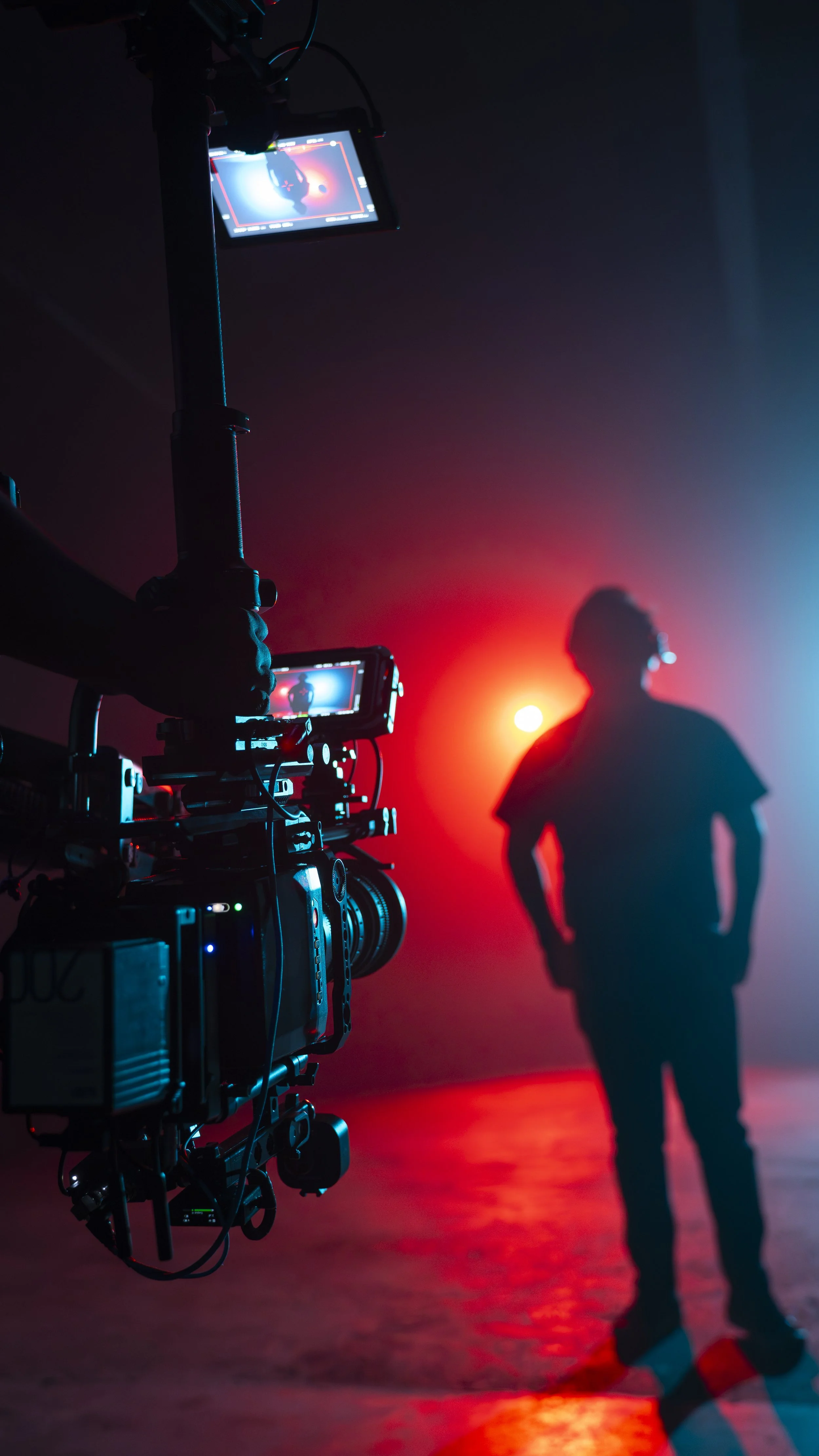A film or photography studio with a person's silhouette in the background, surrounded by red and blue lighting, and a camera setup recording the scene.