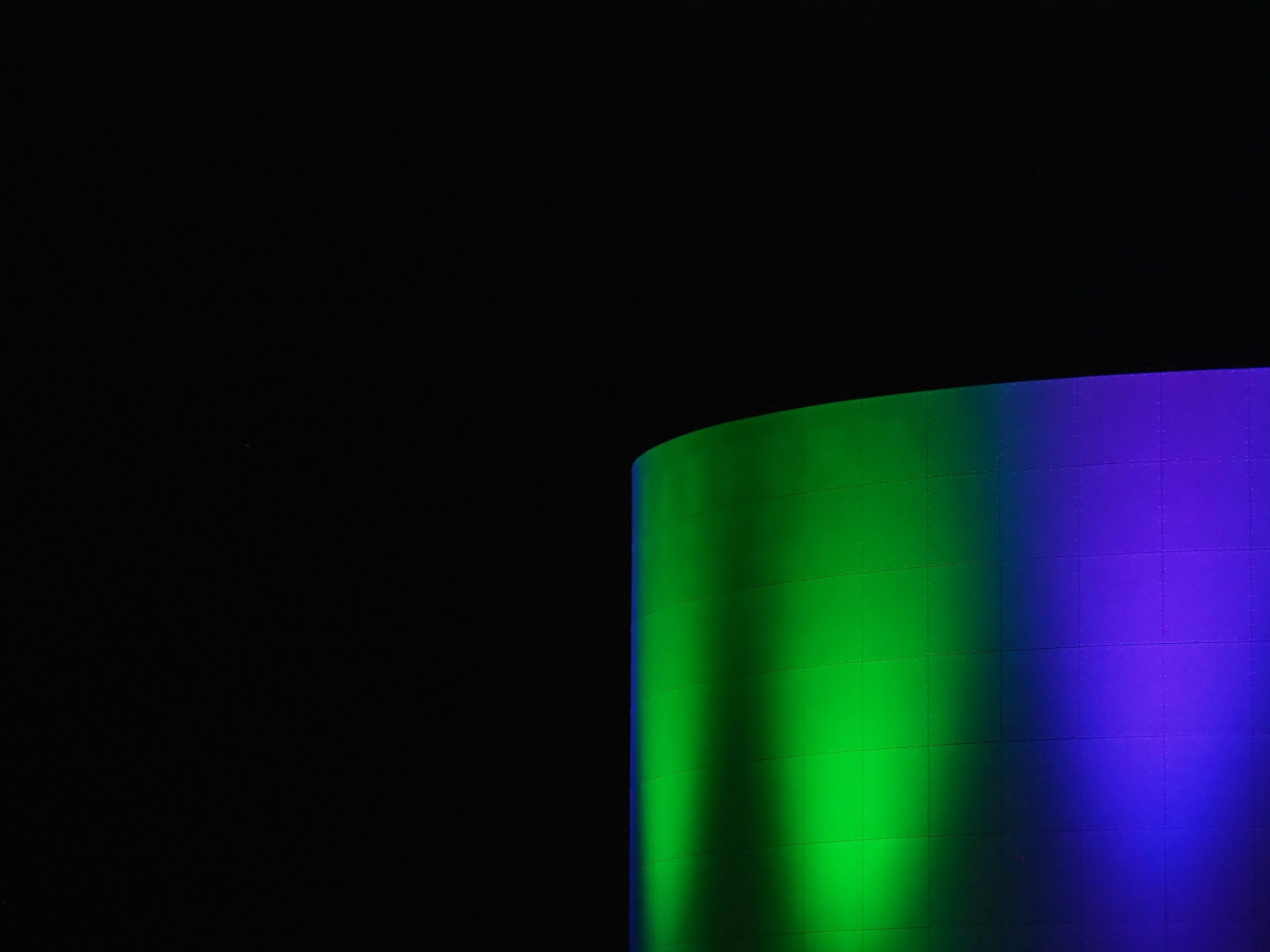 Part of a cylindrical building illuminated with green and purple lights at night, against a dark sky.