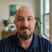 A man with a shaved head and beard wearing a navy polo shirt, sitting indoors near a large window with natural light, plants, and artwork in the background.