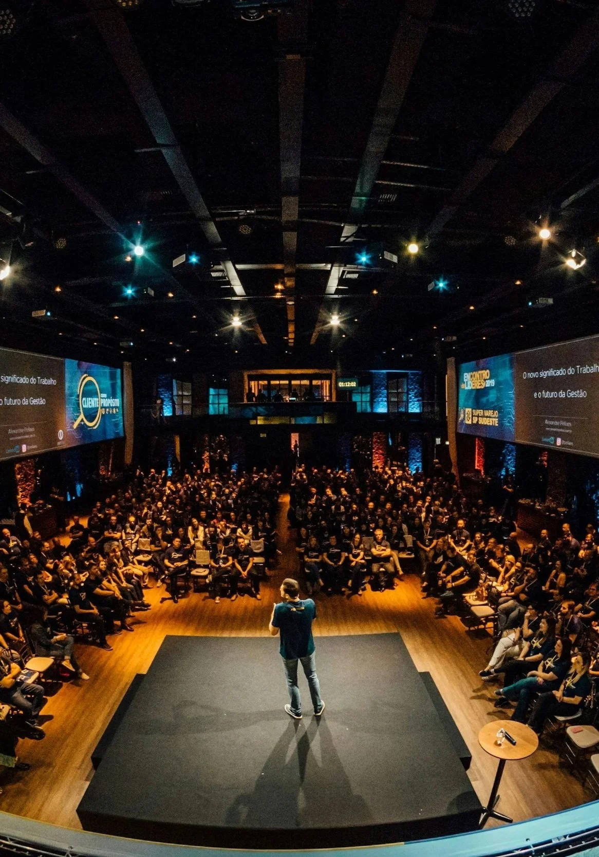 A man stands on a stage in front of a large audience in a conference room, giving a presentation. The room is equipped with multiple large screens displaying text in Portuguese and a mix of warm and cool lighting.