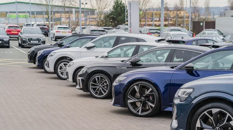 Line of modern cars parked in a dealership lot with a building and trees in the background.