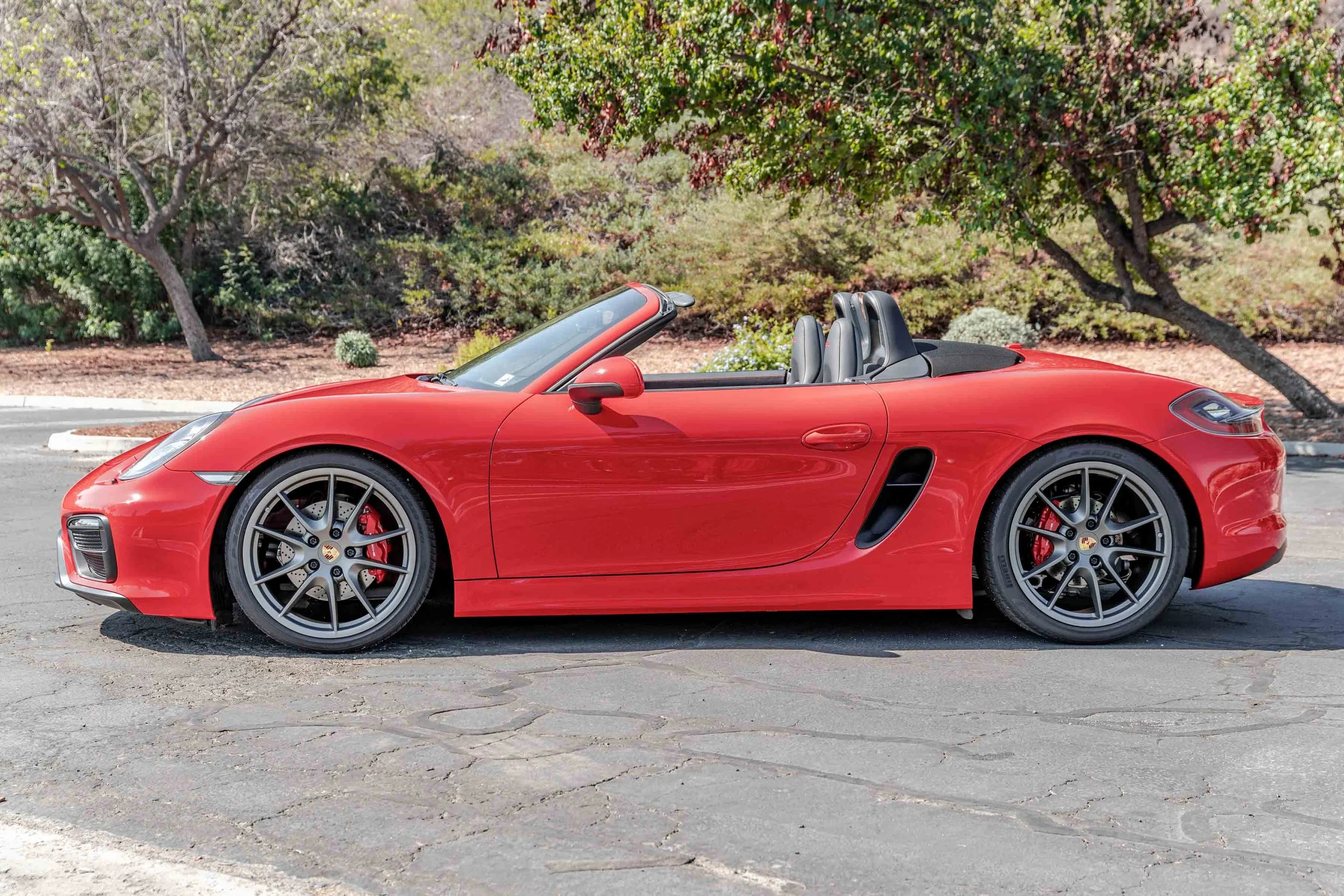 Red sports convertible car parked on a paved area with trees and bushes in the background.