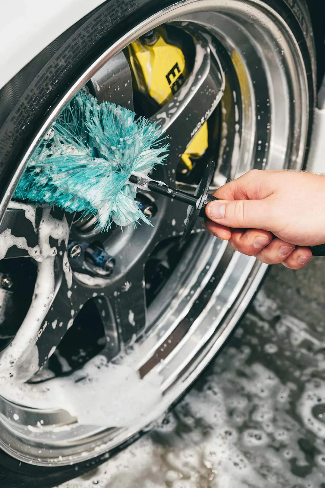 Person hand scrubbing a car wheel with a blue brush and soap suds.