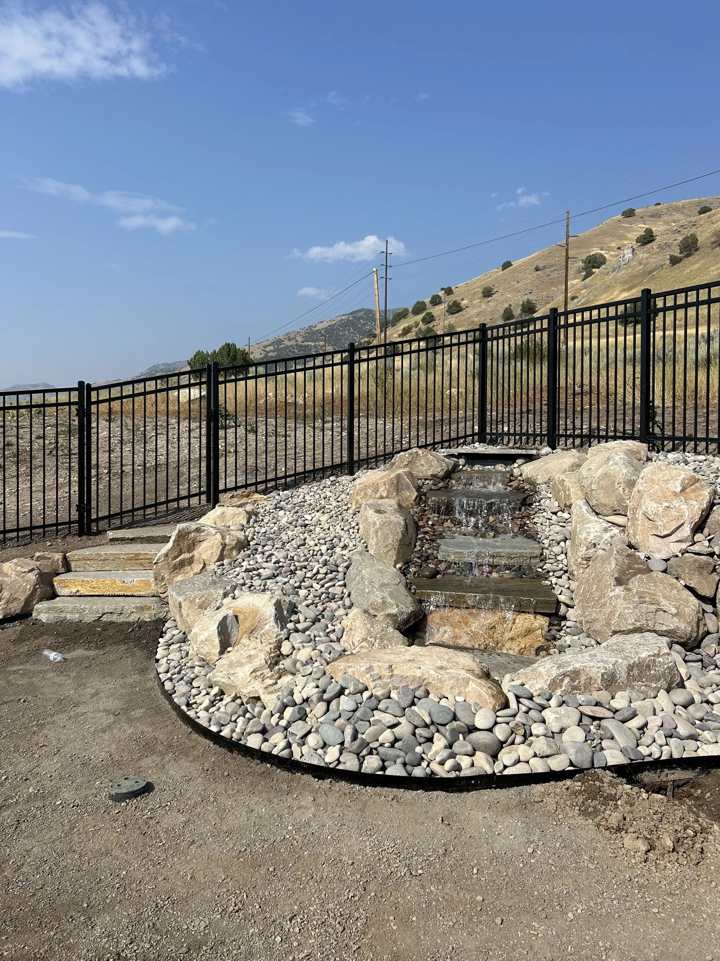A small artificial waterfall with rocks and pebbles, enclosed by a black metal fence, set against a backdrop of hills and a monolithic sky.