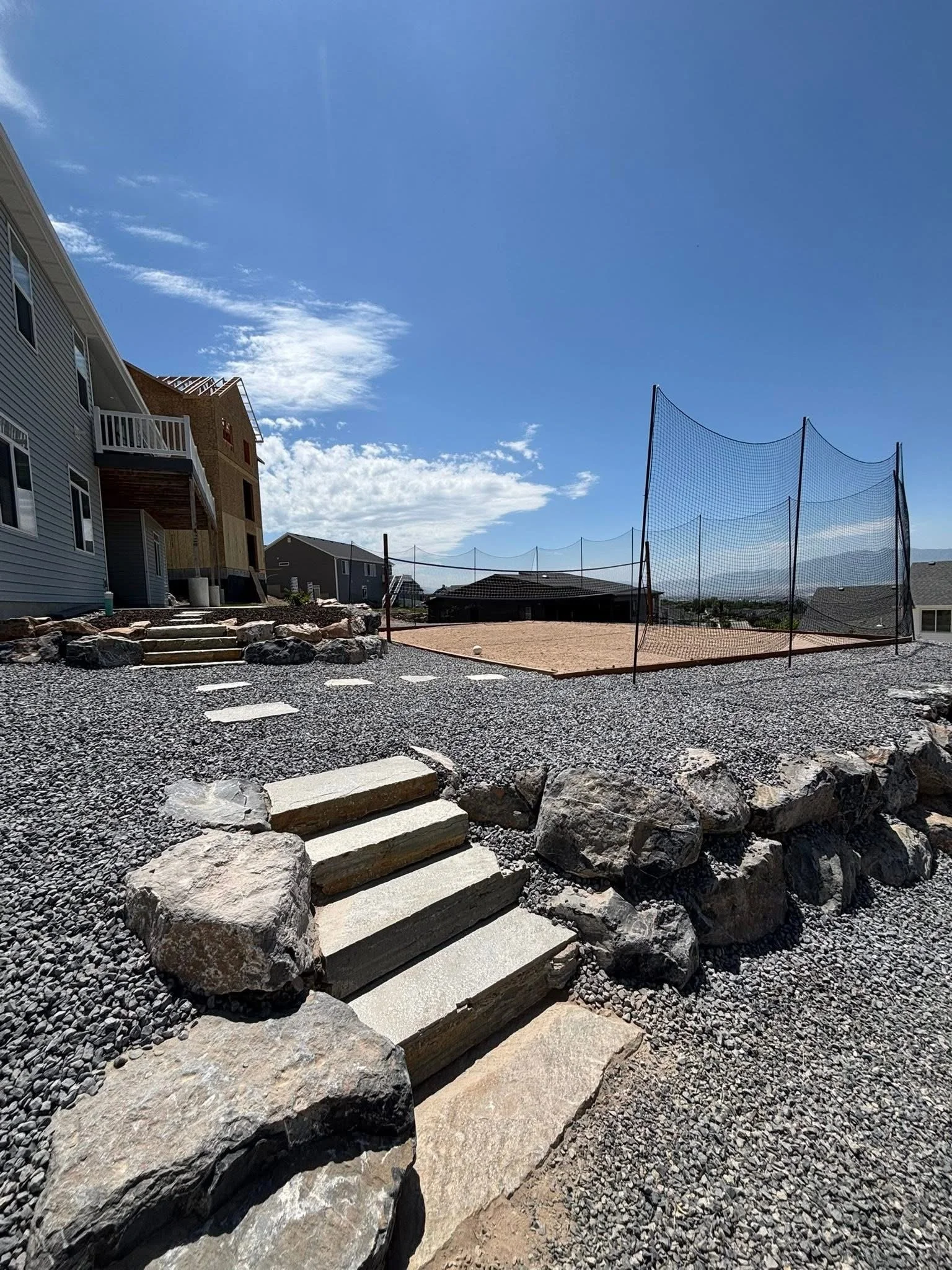 A backyard with gravel ground, stone steps leading to a sand volleyball court enclosed with a tall net, and nearby houses under a blue sky with some clouds.