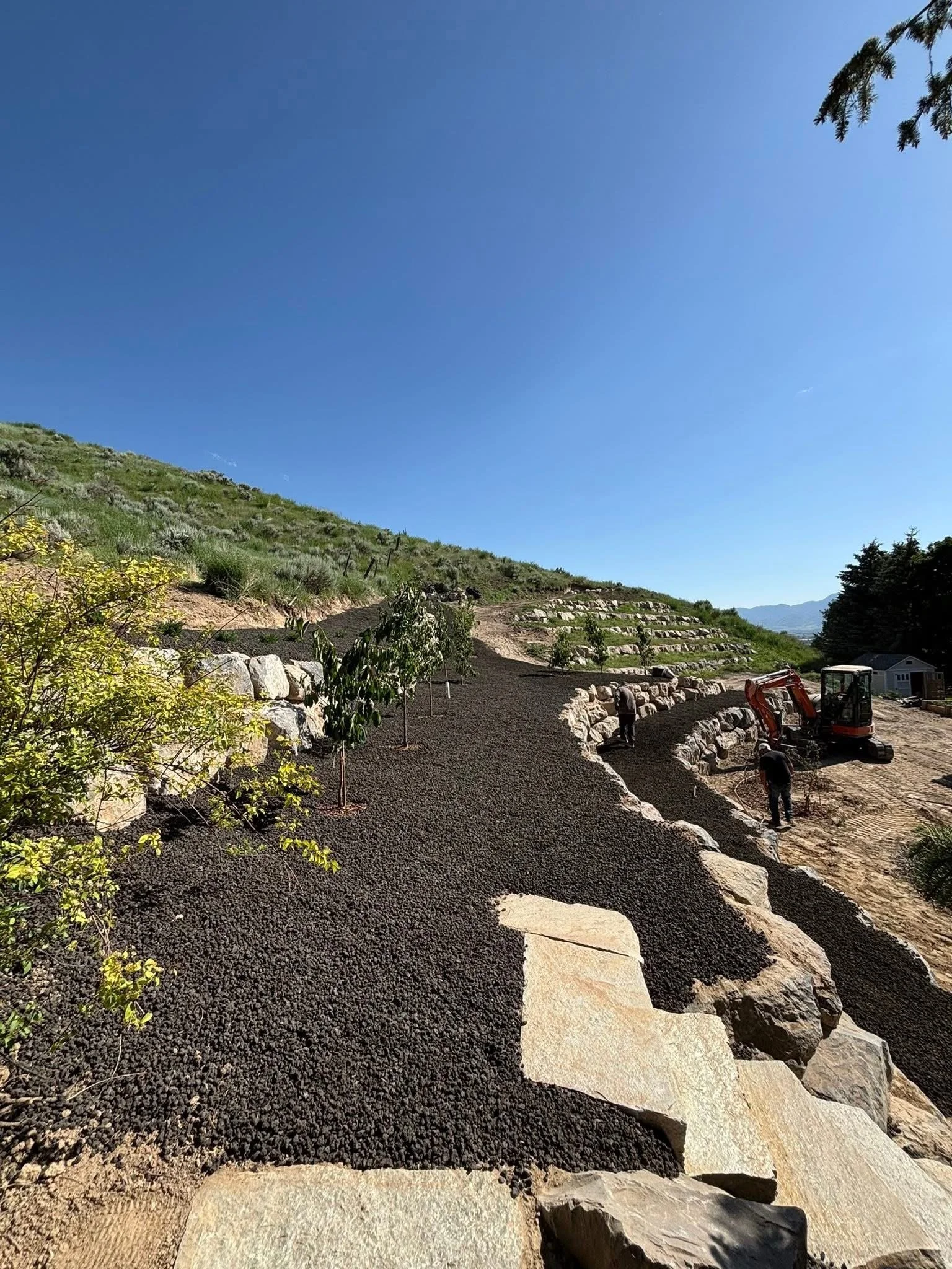 A landscape with a partly cloudy blue sky, a hilly terrain with green vegetation, a dirt pathway being lined with rocks, and construction workers with machinery working on the hillside.