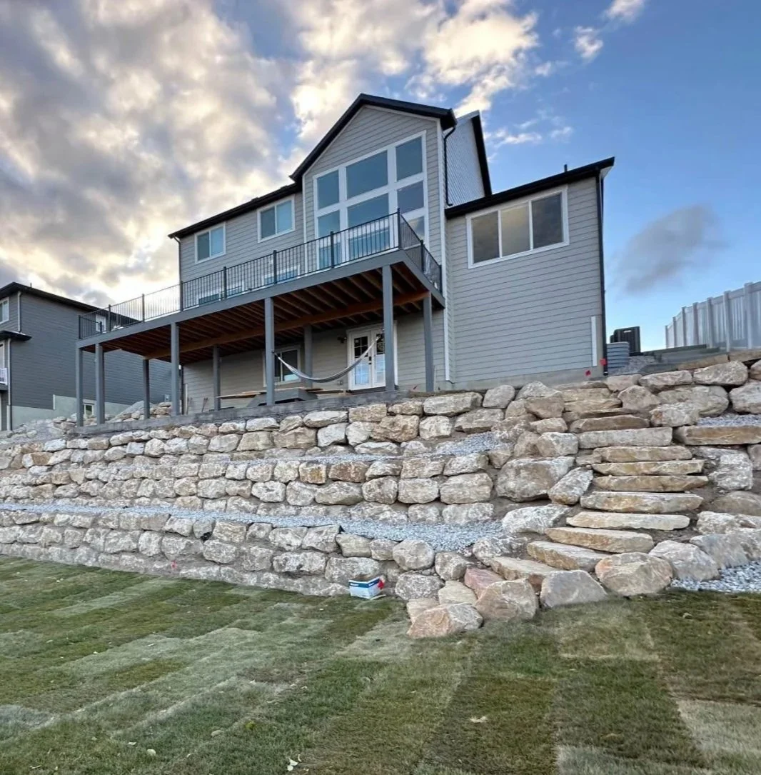 A modern house on a hillside with a stone retaining wall and a wooden deck at the back, under a partly cloudy sky.