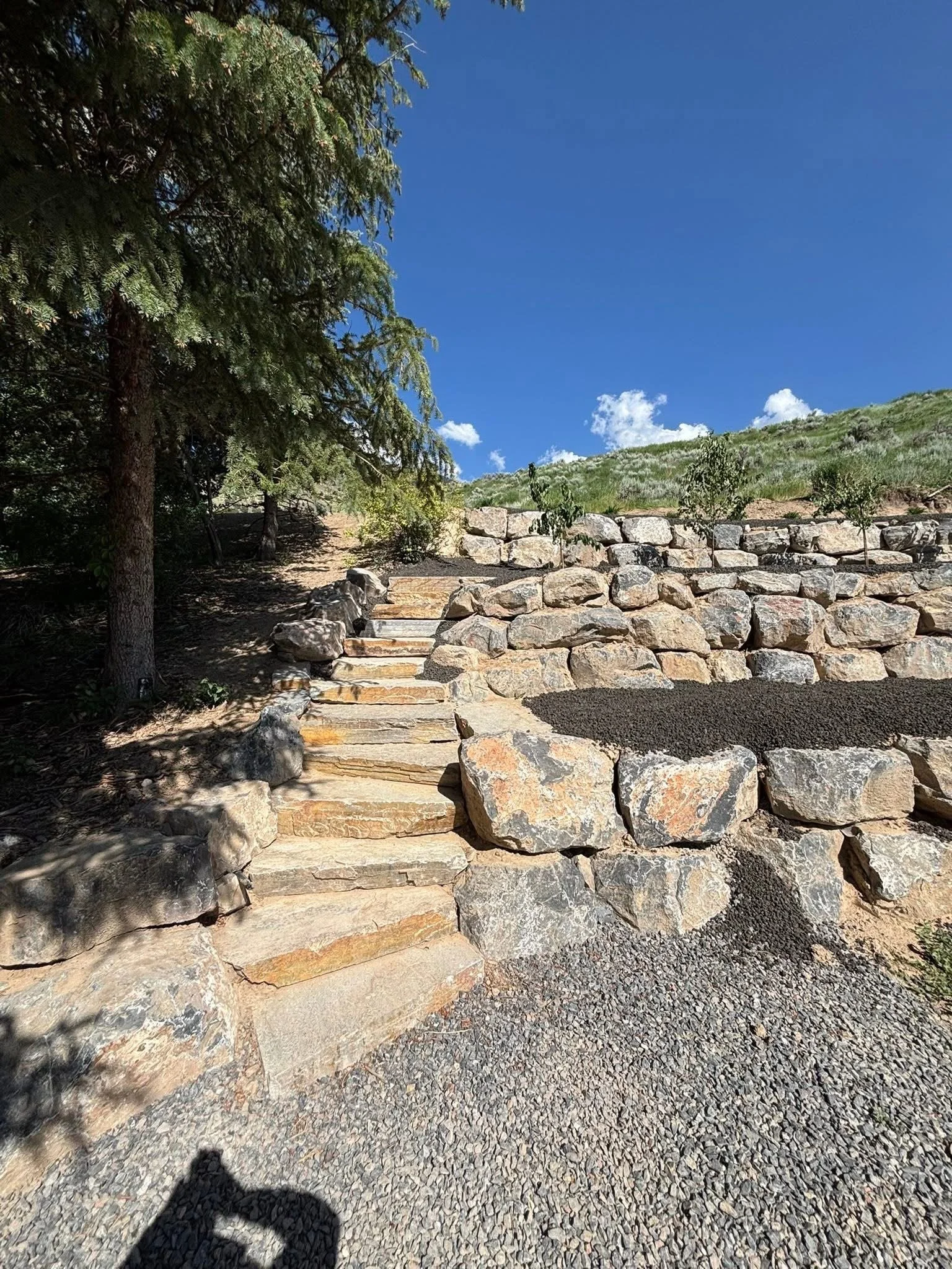 Stone steps leading upward, bordered by large rocks, in a sunny outdoor landscape with trees on the left and a grassy hillside in the background under a clear blue sky.