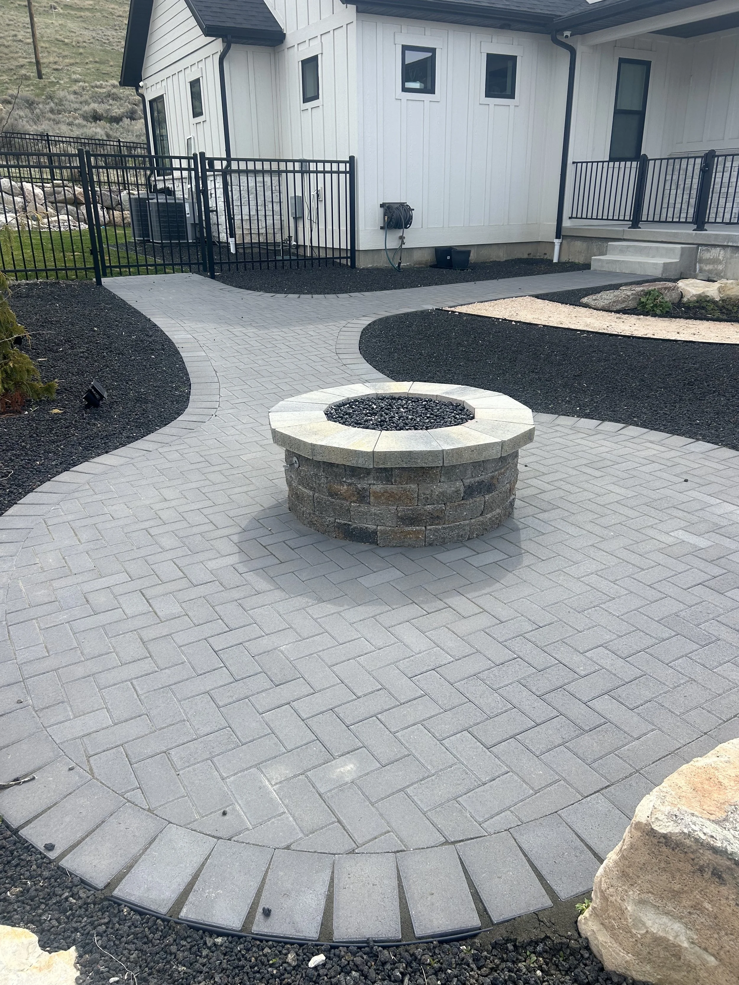 A backyard patio with sidewalk paving, a fire pit, landscaped areas, and a house with a black fence.