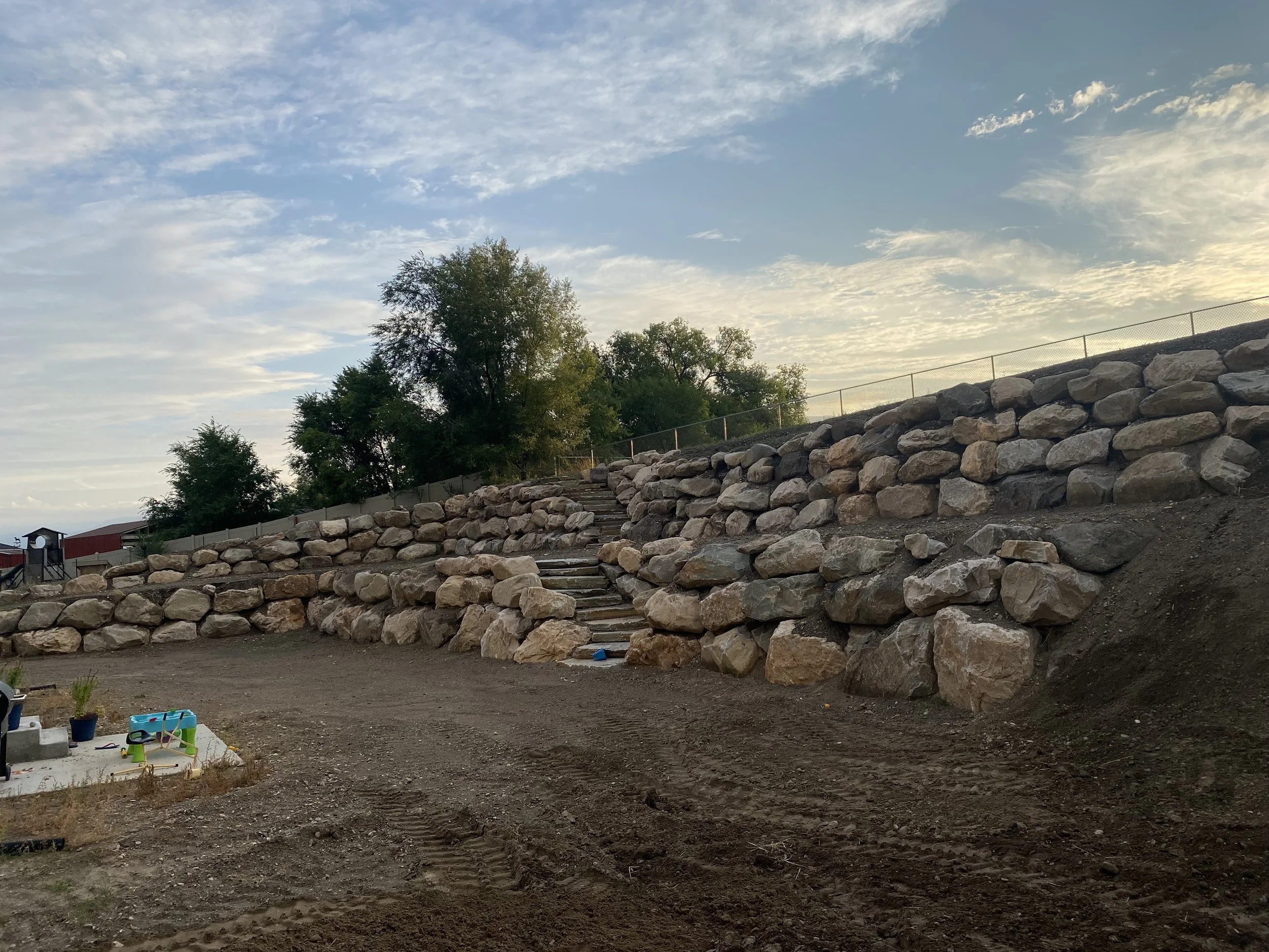 Construction site with a rock retaining wall, dirt ground, a few trees, and a cloudy sky.