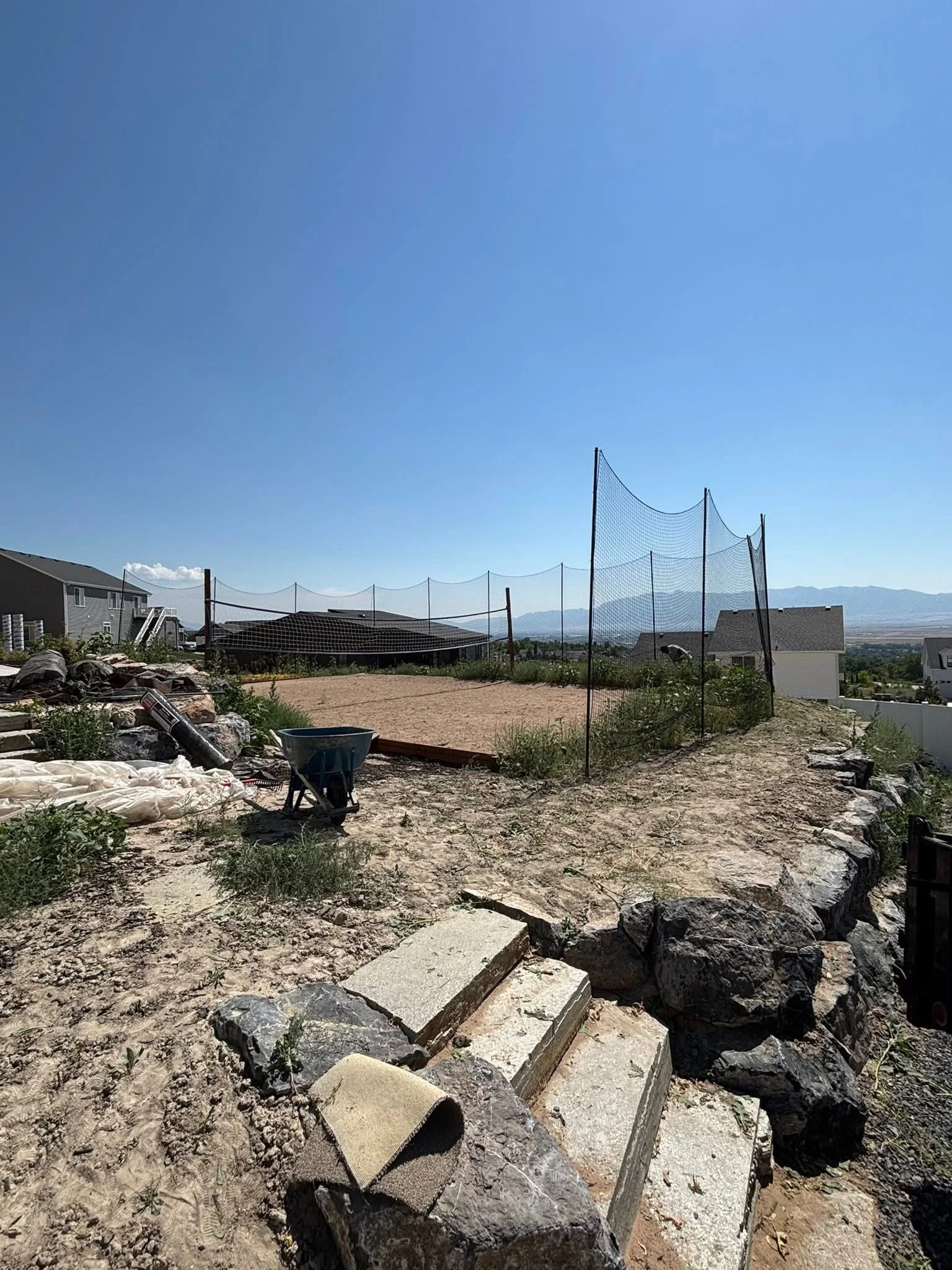 A backyard with a sandy volleyball court enclosed by a tall net, with rocky terrain and green plants in the foreground, and residential houses and mountains in the distance under a clear blue sky.