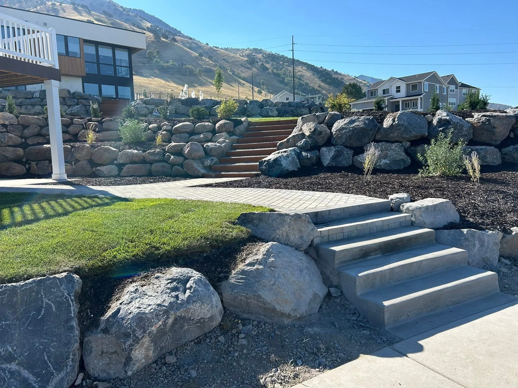 Residential backyard with concrete steps leading up to a rock retaining wall, landscaped with plants and mulch, alongside a paved pathway and a grassy area, under clear blue sky.