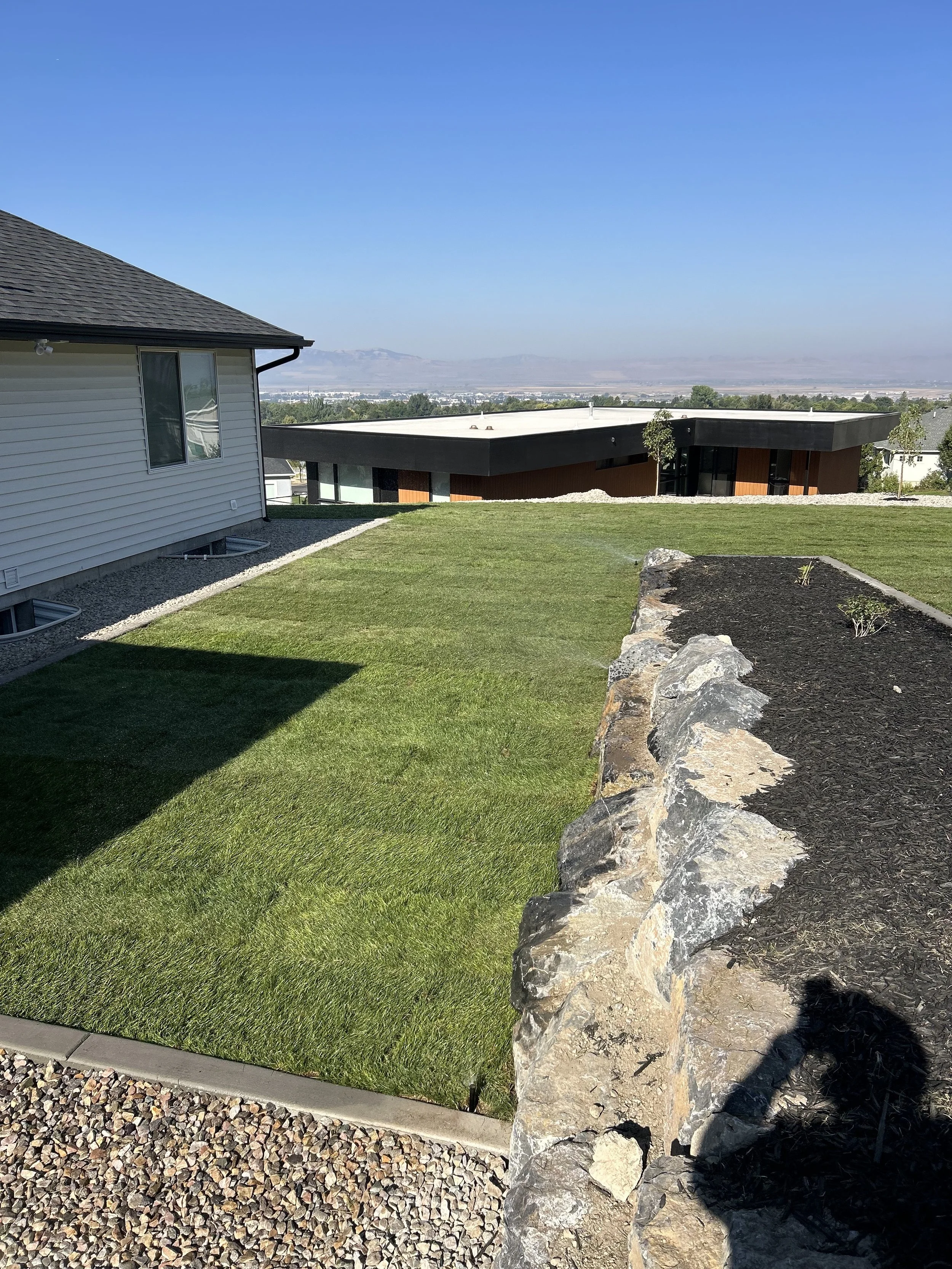 A backyard with a well-maintained green lawn, a white house on the left, a stone border on the right, and modern houses and mountains in the background under a clear blue sky.