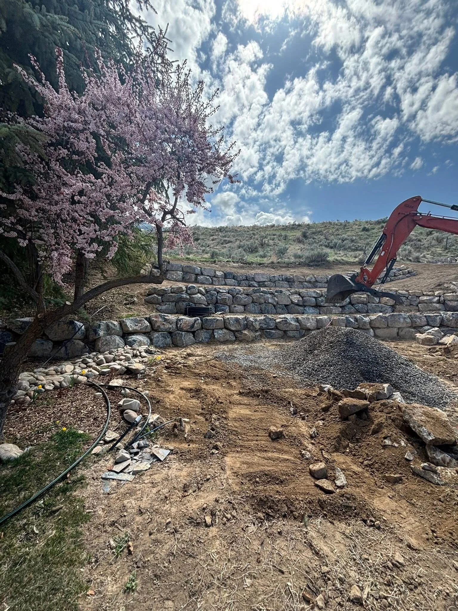 Construction site with a partially built stone wall, an excavator, dirt, rocks, and a flowering tree with pink blossoms under a partly cloudy sky.