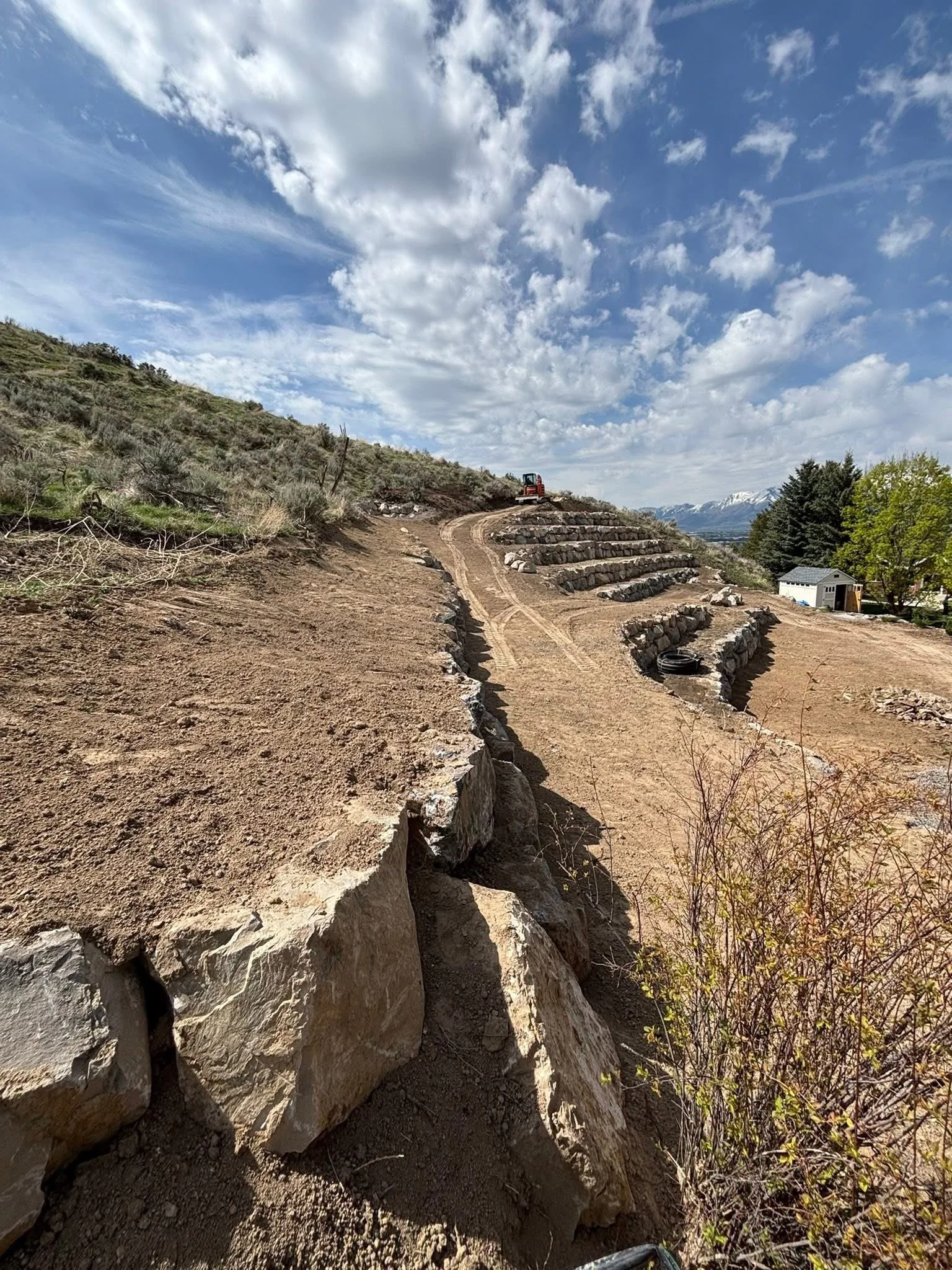 hillside terraced with large stones, a dirt path winding uphill, construction equipment at the top, green trees, and snow-capped mountains in the background under a partly cloudy sky.