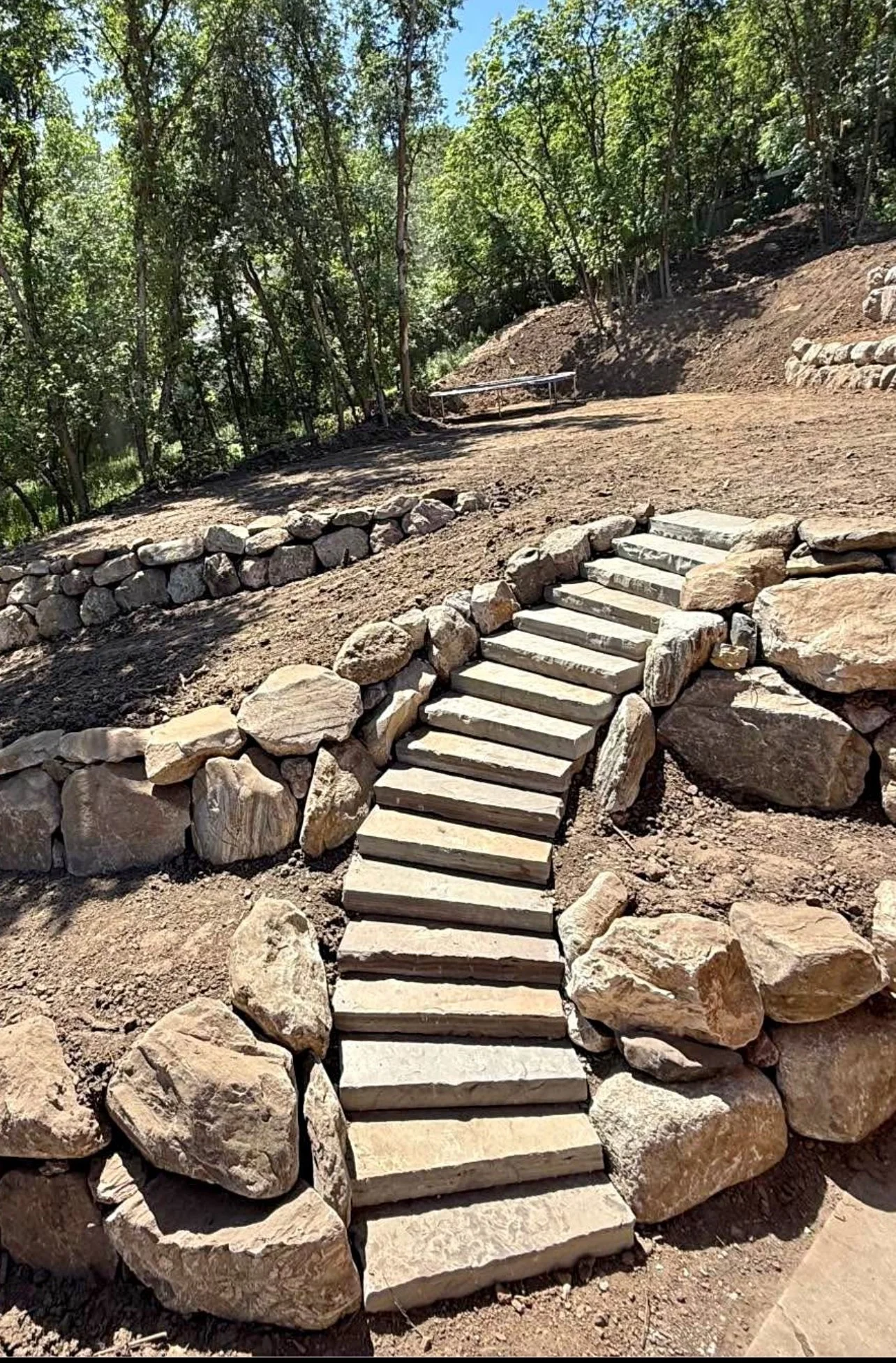 Stone staircase with large rocks on sides in a wooded area, under construction.