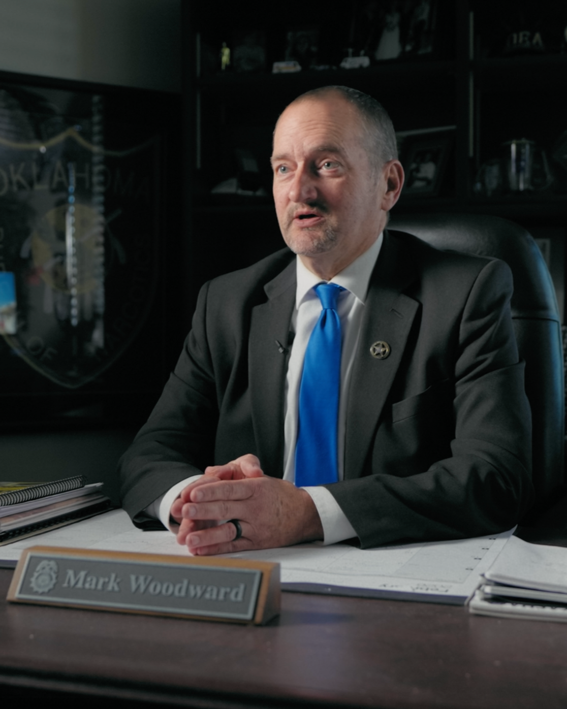 A man in a black suit with a blue tie sitting at a desk, with a nameplate that reads "Mark Woodward." He is in an office with dark wood furniture and shelves containing framed photographs and decorative items.