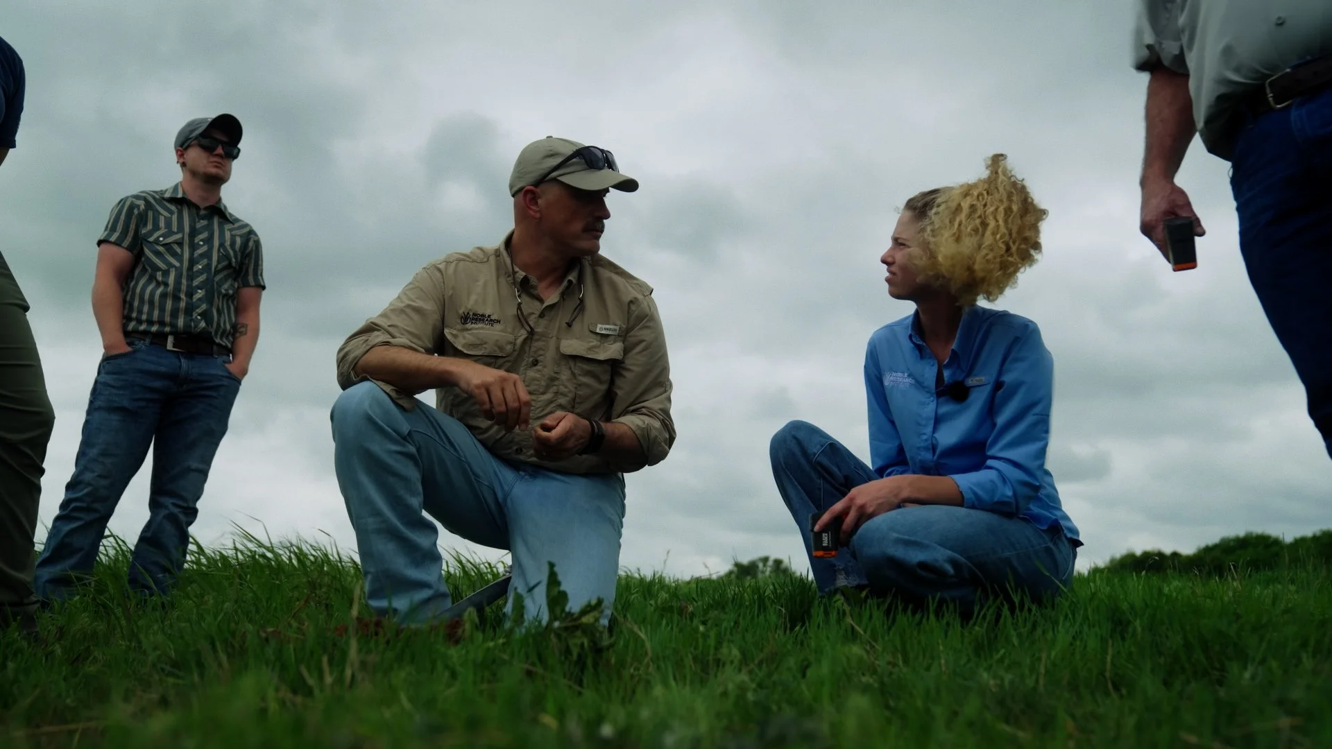 Group of five people outdoors on grassy field, two sitting and three standing, engaged in conversation, under cloudy sky, mostly focused on a woman with curly blonde hair and a man in khaki shirt.